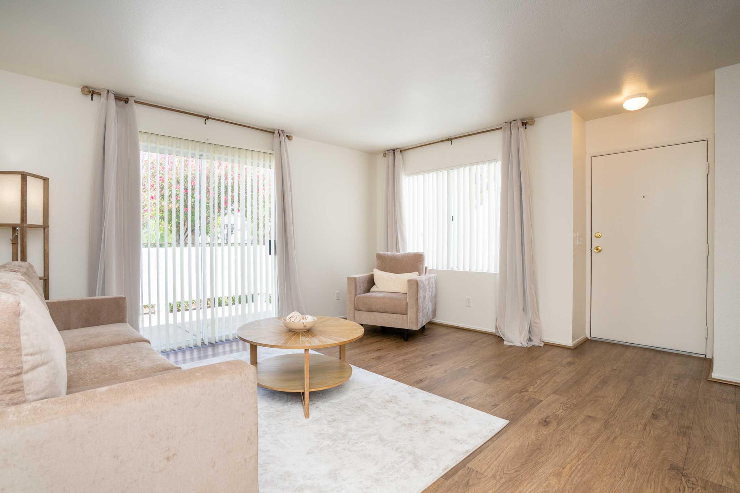 A bright, minimalist living room featuring a light beige sofa and an armchair, a round wooden coffee table, and a light-colored area rug. Two large windows with vertical blinds let in natural light, and soft curtains frame the windows. The entrance door is visible on the right.
