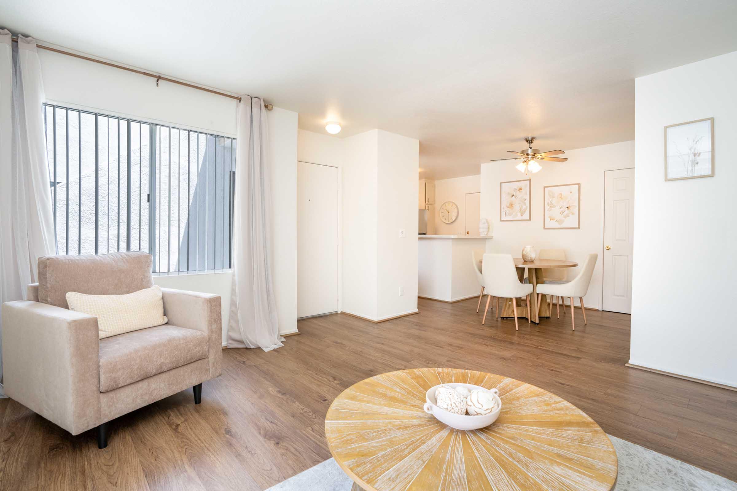 A spacious living area featuring light wood flooring, a beige armchair with a cushion, and a round table with chairs. Large windows with curtains allow natural light, and a clock and framed artwork decorate the walls. The kitchen area is visible in the background.
