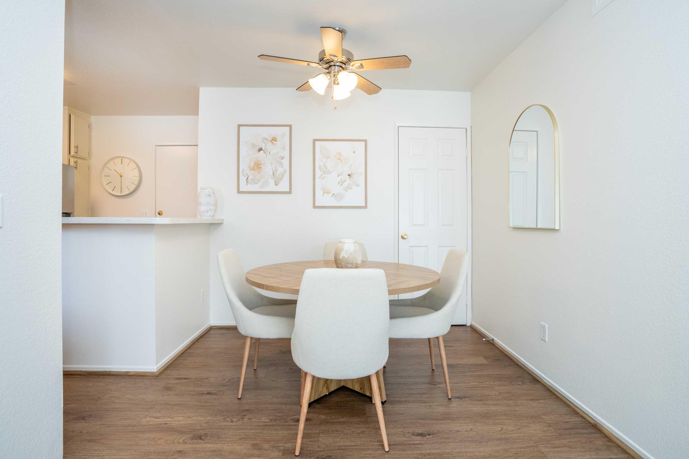 Modern dining area featuring a round wooden table with light-colored upholstered chairs. A ceiling fan is overhead, and soft white walls create a bright atmosphere. Two framed floral artworks hang on the wall, complemented by a mirror. The space is open with a view into the adjacent kitchen area.