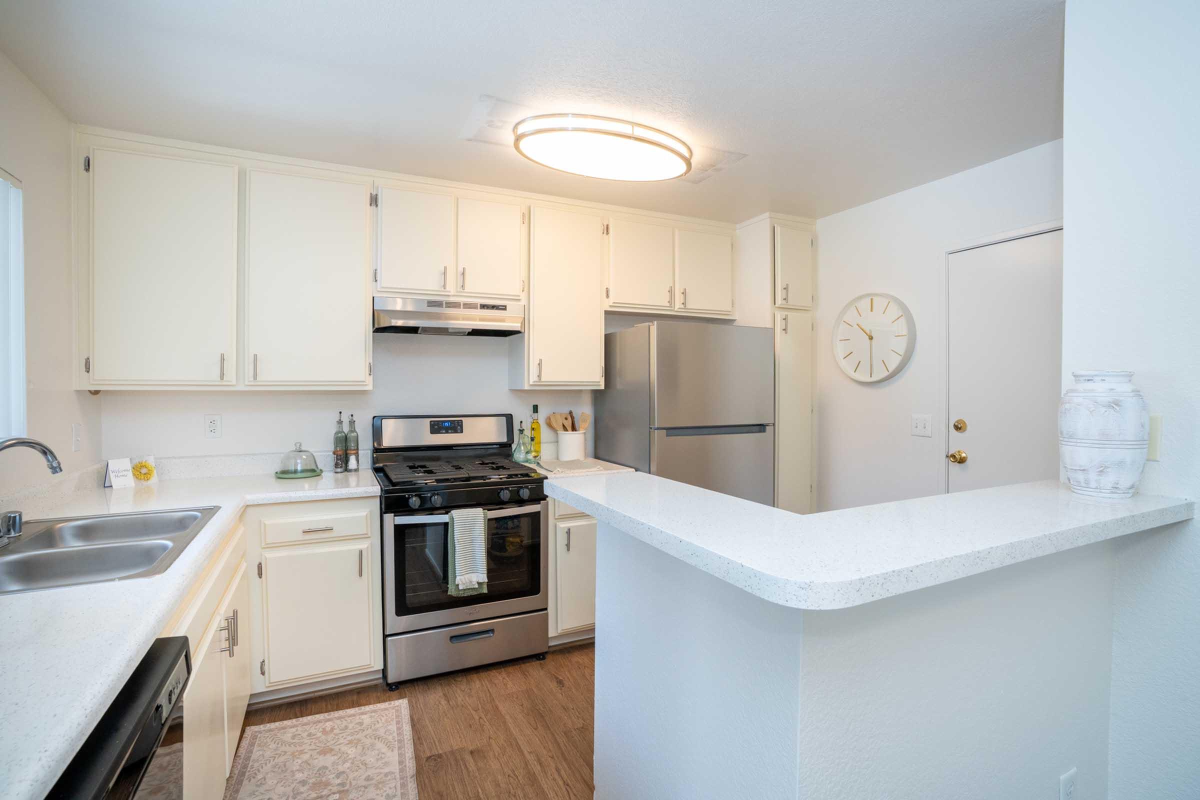 Bright and modern kitchen featuring white cabinets, stainless steel appliances, and a spacious countertop. A large circular light fixture illuminates the area, while a clock adorns the wall. The setup includes a double sink and a cozy rug on the floor, creating a welcoming atmosphere.