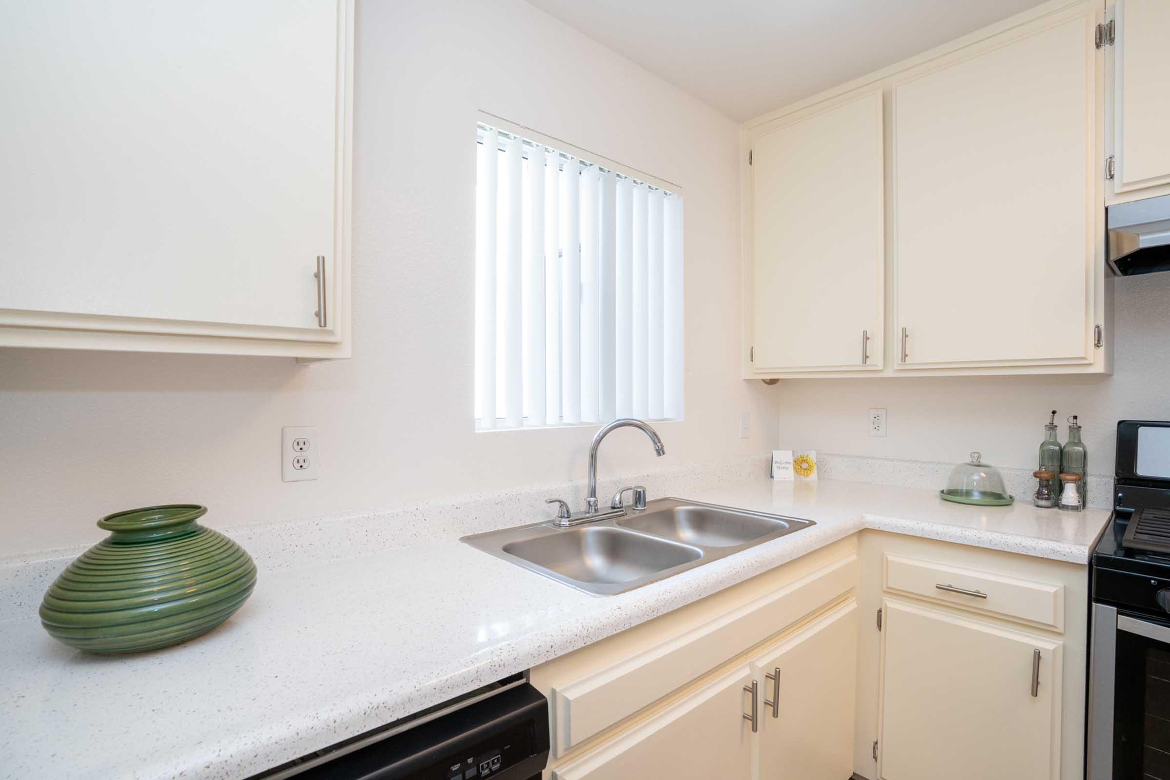 Bright kitchen featuring a two-basin stainless steel sink, light-colored cabinets, and a countertop made of a white speckled material. A green decorative vase sits on the counter, and a window with vertical blinds allows natural light to illuminate the space. Modern appliances and organized decor complete the inviting atmosphere.