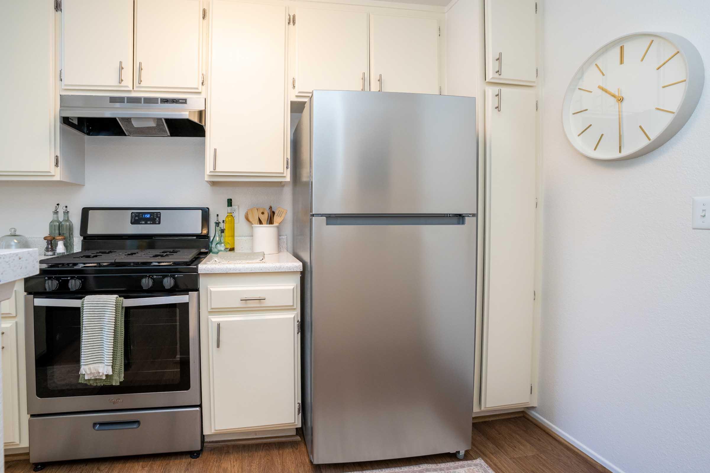 A modern kitchen featuring a silver refrigerator beside a black stove with an oven. The cabinets are light-colored, and a wall clock with a minimalist design is mounted nearby. The countertop is clean, adorned with utensils and a bottle, reflecting a tidy and functional cooking space.
