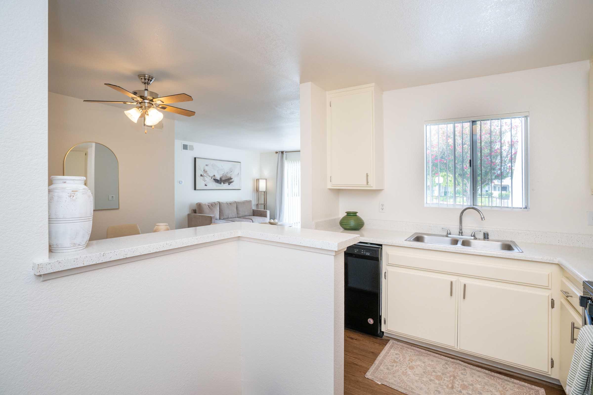 A well-lit kitchen featuring white cabinets, a double sink, and a window with blinds. A beige rug is placed in front of the sink. In the background, a living area is partially visible with a beige couch and decorative wall art. A ceiling fan is installed in the kitchen area.