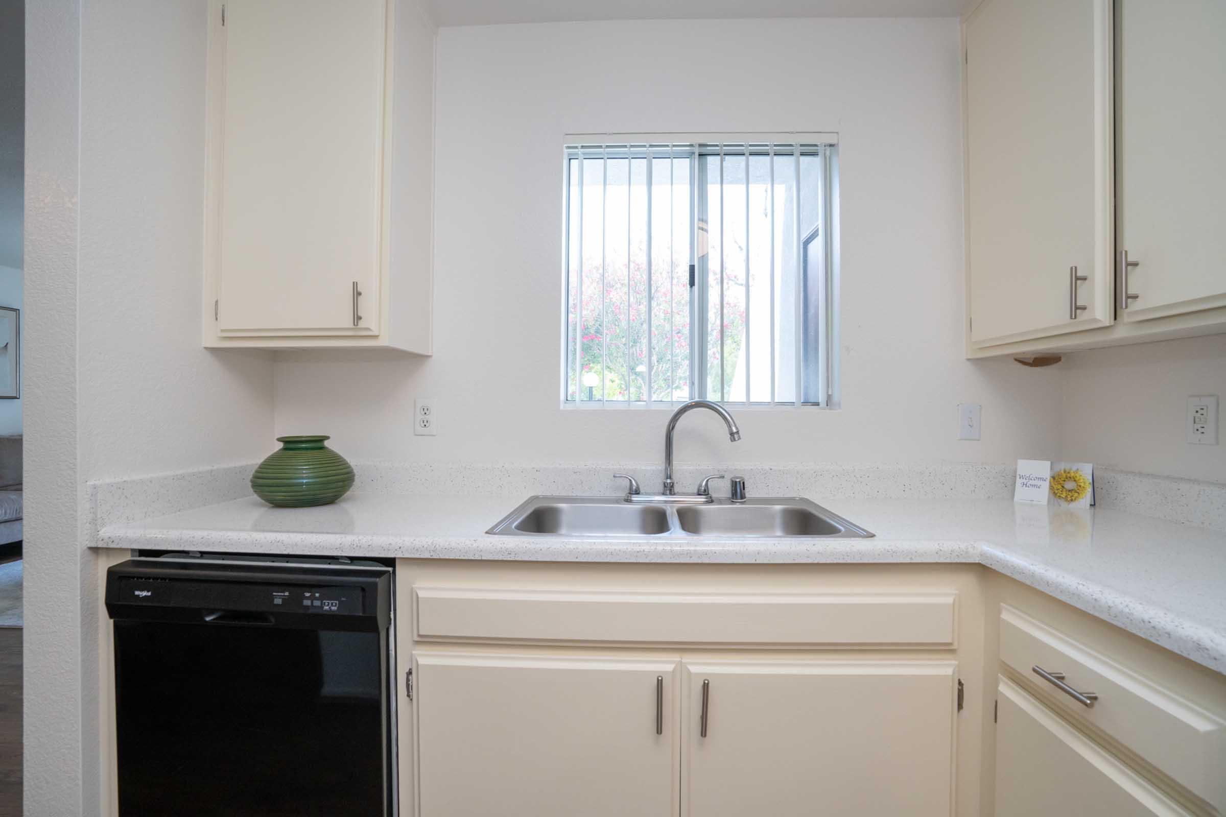 Modern kitchen with light-colored cabinets, a double sink, and a small green decorative vase. The countertop is made of speckled material, and there's a window above the sink letting in natural light. A black dishwasher is visible on the left side of the sink area.