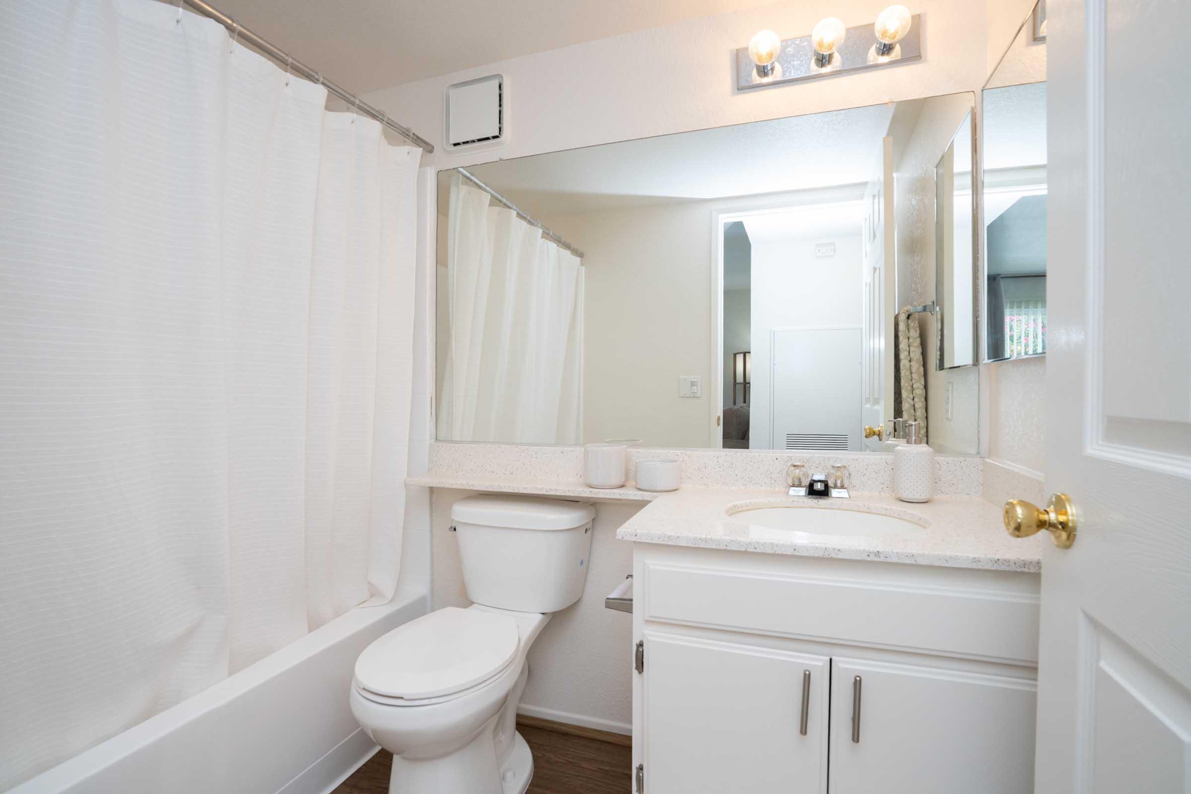 A clean, modern bathroom featuring a white shower curtain, a toilet, and a vanity with a mirror. The countertop is adorned with decorative items, and the lighting is provided by overhead fixtures. The walls are painted light colors, creating a bright and spacious feel.