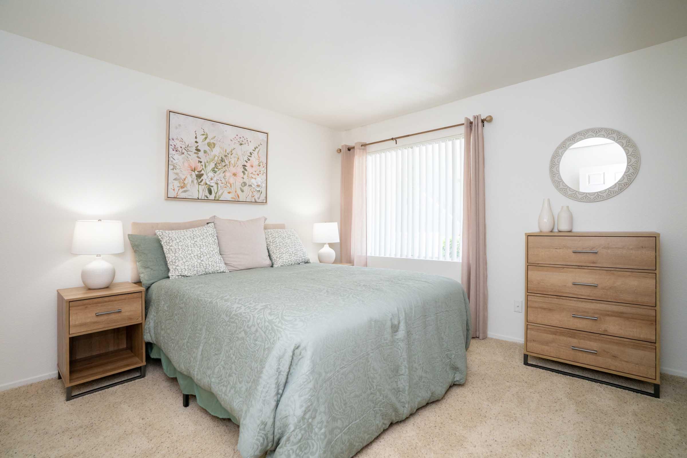 A bright and inviting bedroom featuring a queen-sized bed with a light green blanket, complemented by decorative pillows. There are two bedside lamps, a wooden dresser, and a round mirror on the wall. A window with sheer curtains allows natural light to enter, enhancing the cozy atmosphere.