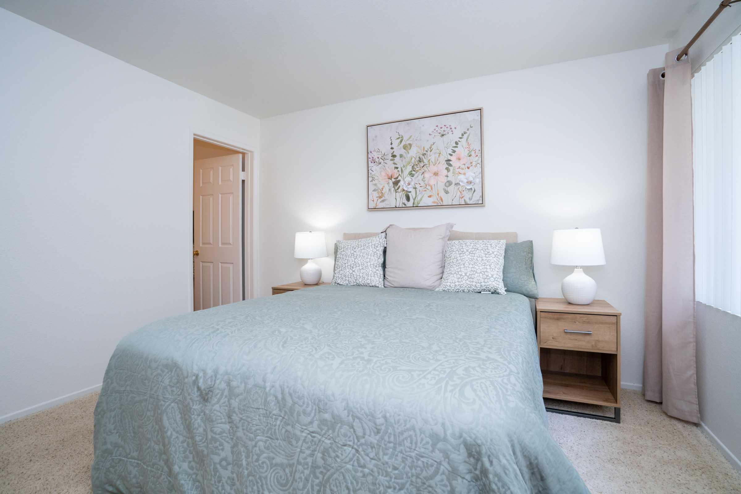 A cozy bedroom featuring a large bed with a light teal covering, two bedside tables with white lamps, and a floral wall art above the bed. The room has neutral walls and carpet, with a window letting in natural light and a door leading to another room.
