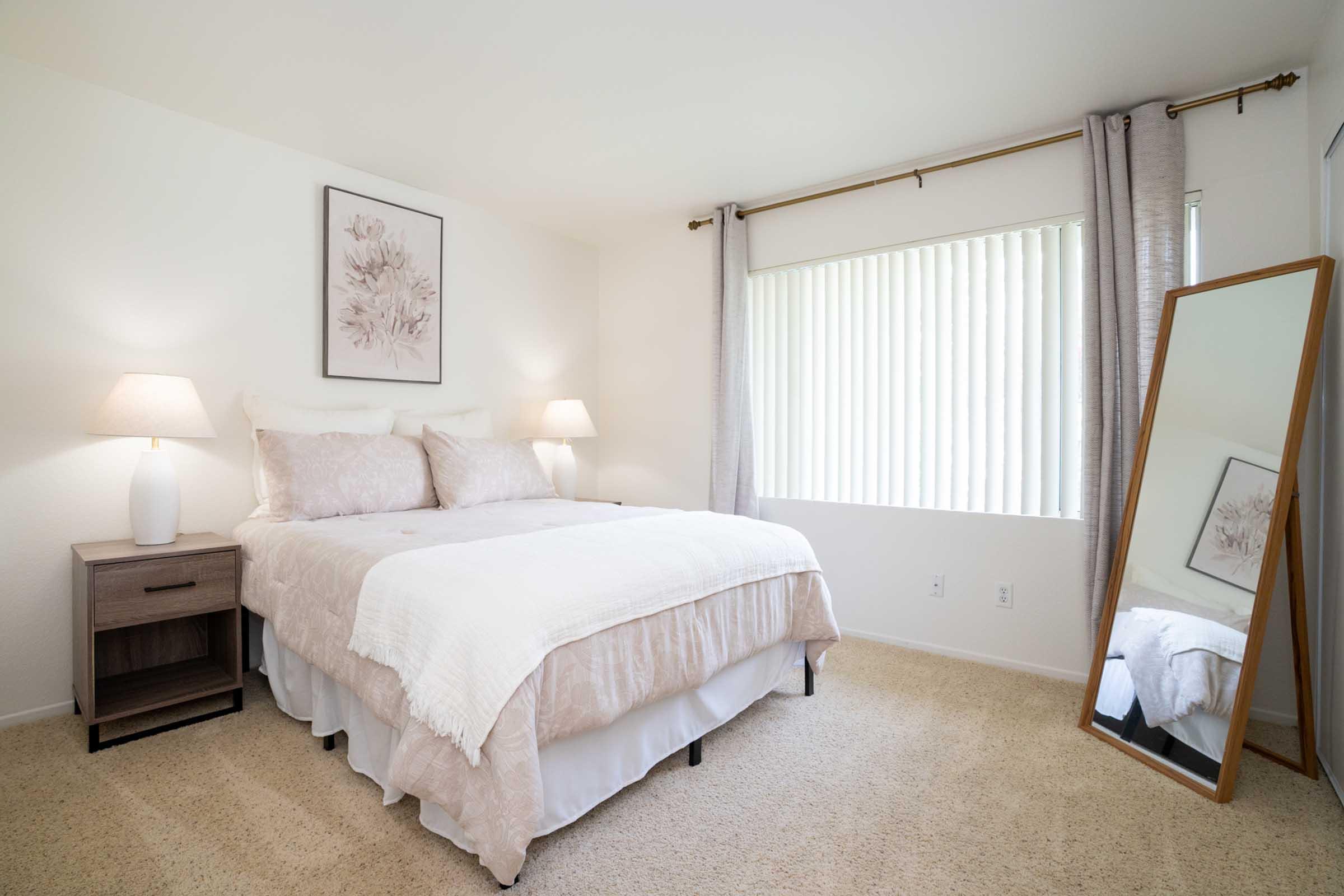 A cozy bedroom featuring a bed with light-colored linens and decorative pillows, a nightstand with a lamp, sheer window curtains, and a large mirror. The walls are painted in soft tones, and a piece of floral artwork decorates one wall, creating a serene and inviting atmosphere.