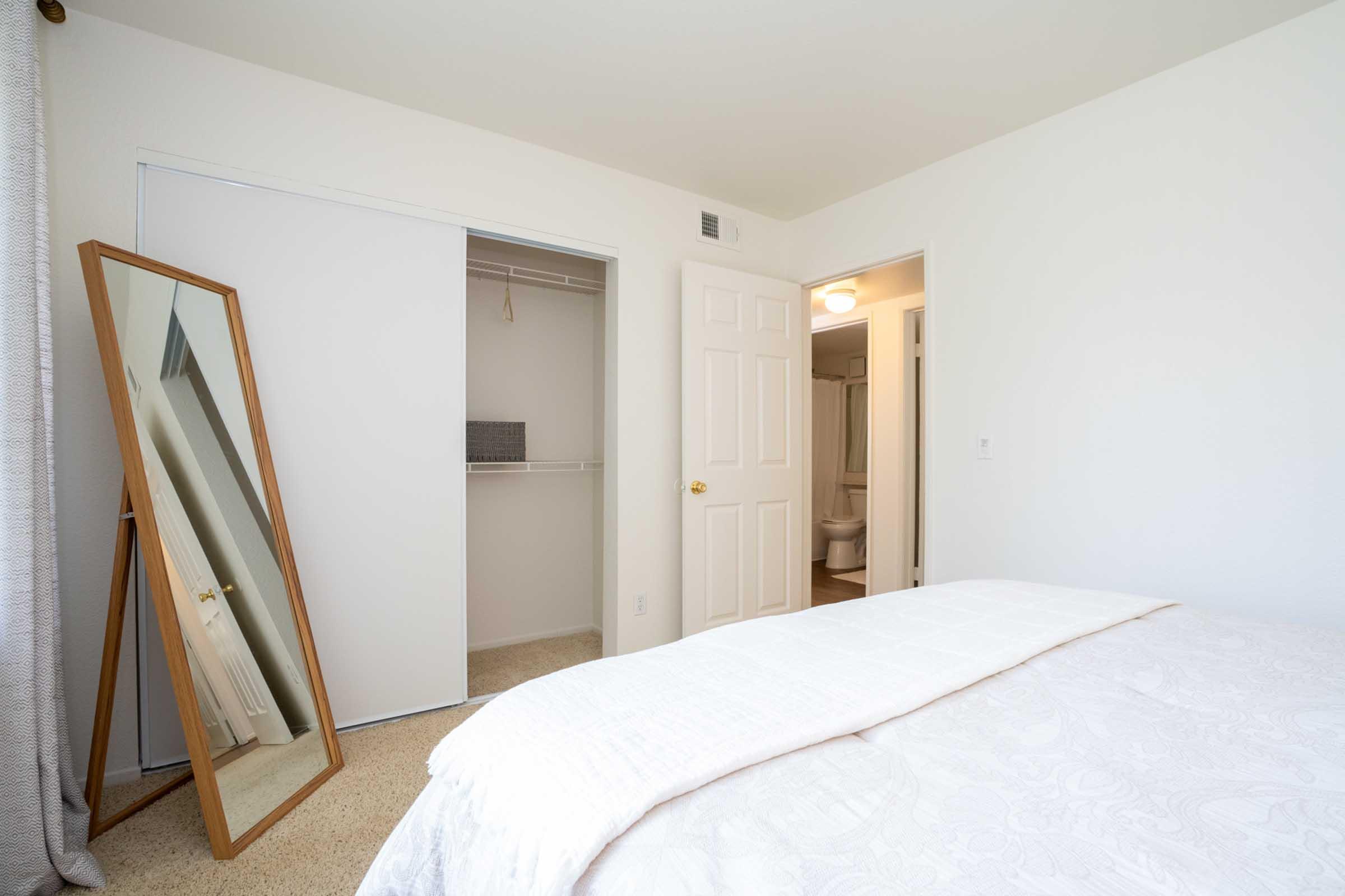 A well-lit bedroom featuring a neatly made bed with a white comforter. A large, leaning mirror is positioned against the wall, reflecting the room. There’s a closet door on the left, slightly open, and a bathroom door is visible in the background. The walls are painted white, creating a clean and spacious atmosphere.