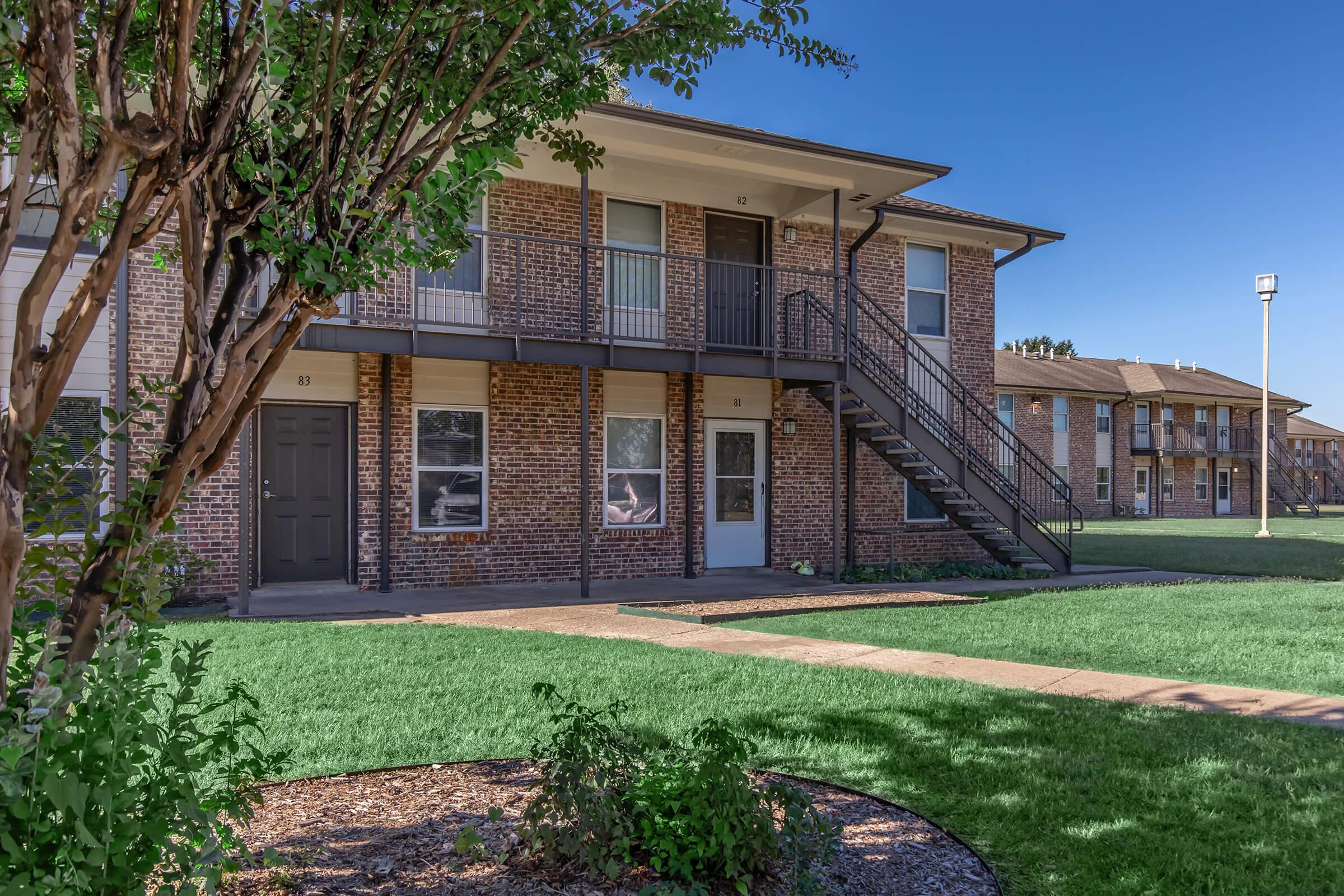 A residential apartment complex featuring two buildings surrounded by well-maintained grass and landscaping. The foreground includes a tree and a walkway leading to the entrances, with stairs leading to the upper floors of the apartments. The sky is clear and blue.