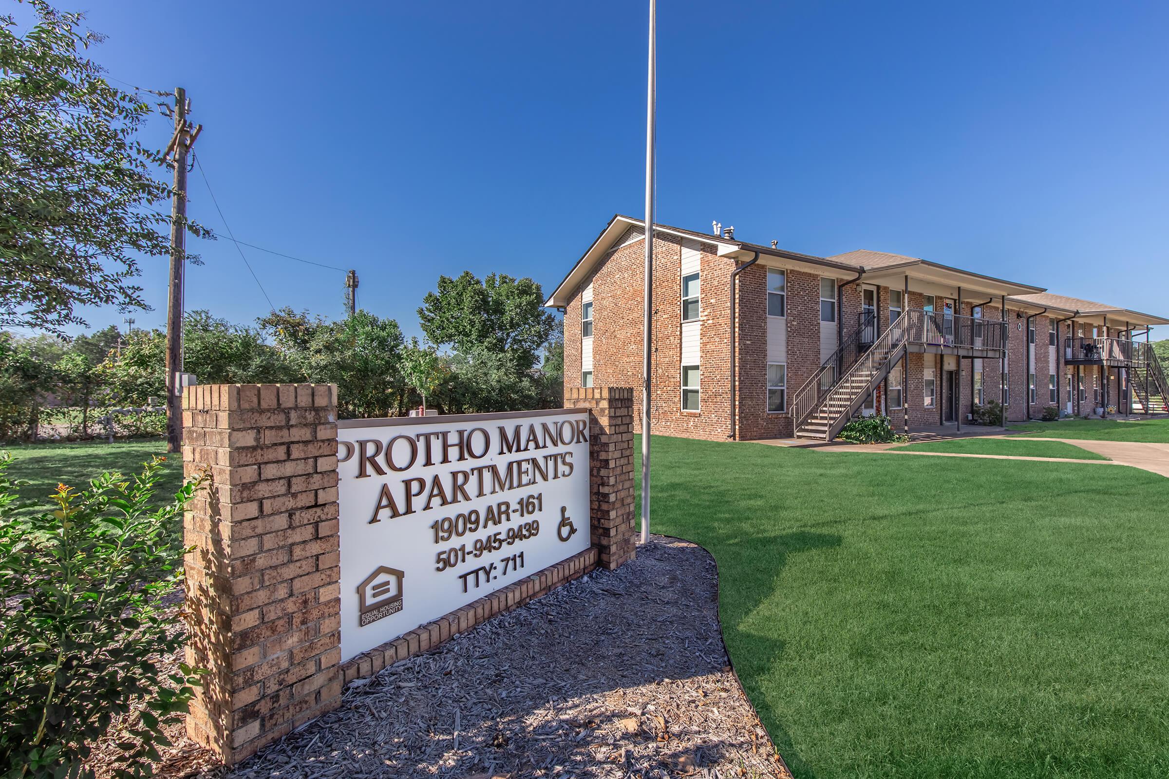 Sign for Protho Manor Apartments, with the address 1909 AR-161. It's a two-story brick apartment building surrounded by green grass, with a clear blue sky above. The sign indicates contact details and accessibility features.
