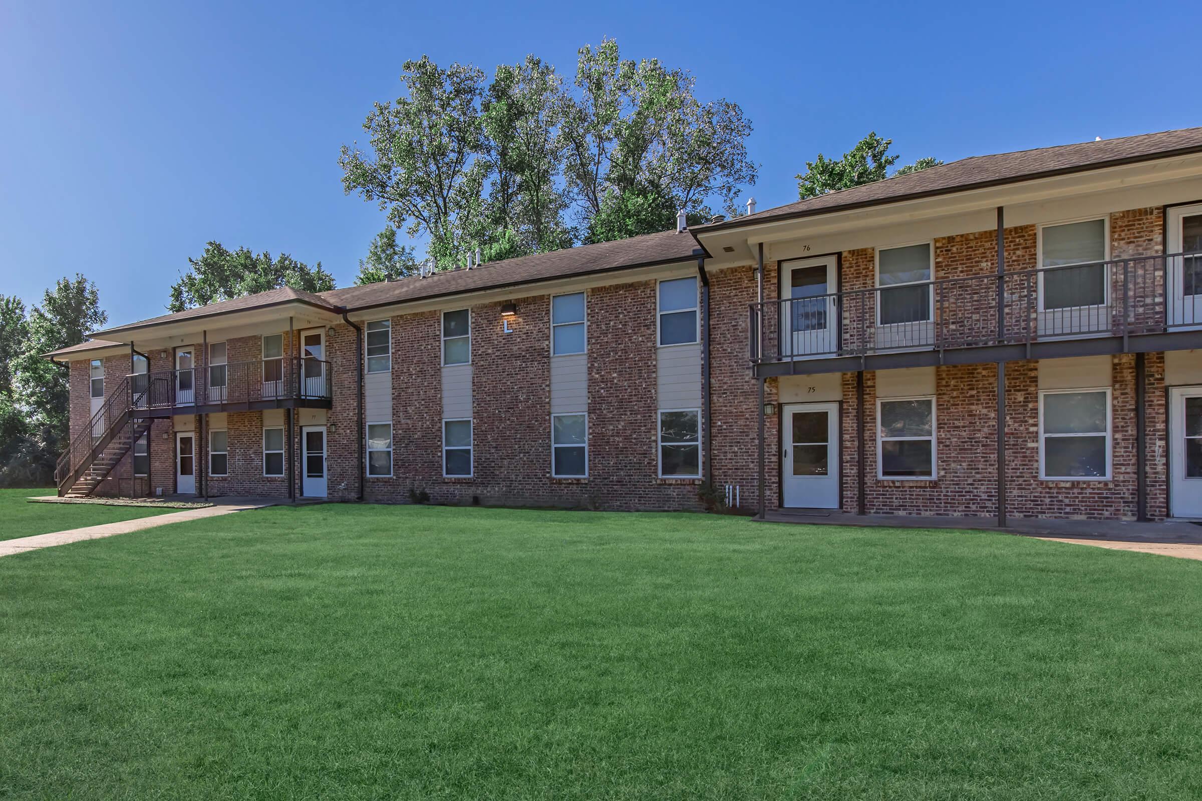 A brick apartment building with multiple units, featuring two levels, balconies, and a well-maintained lawn. Trees in the background and a clear blue sky enhance the scene, making it look inviting and pleasant. The pathways lead to the entrance of each apartment.