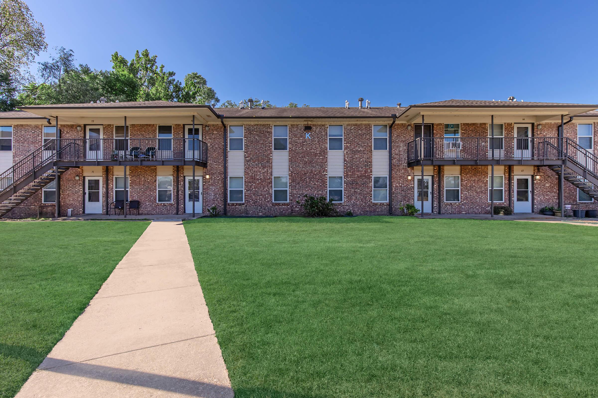 Two-story brick apartment building with balconies on the upper level, surrounded by a well-maintained grassy area. A paved walkway leads through the lawn, connecting the entrance to the apartments. Clear blue sky overhead, creating an inviting atmosphere.
