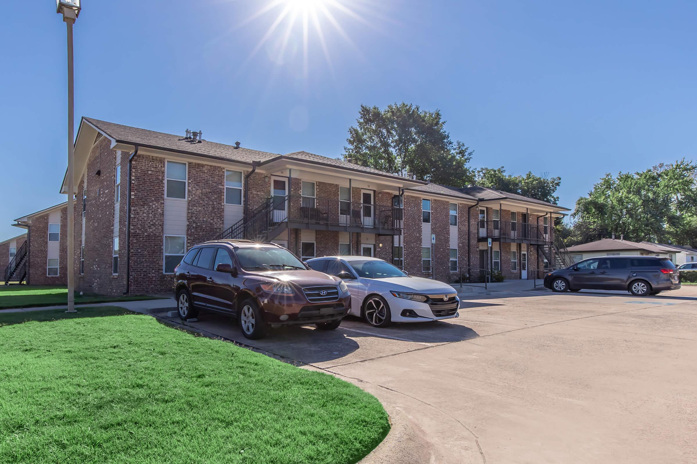 A view of a multi-unit residential building with brick exterior on a sunny day. The scene includes several parked cars in front of the building, surrounded by green grass and trees, with the sun shining brightly overhead.