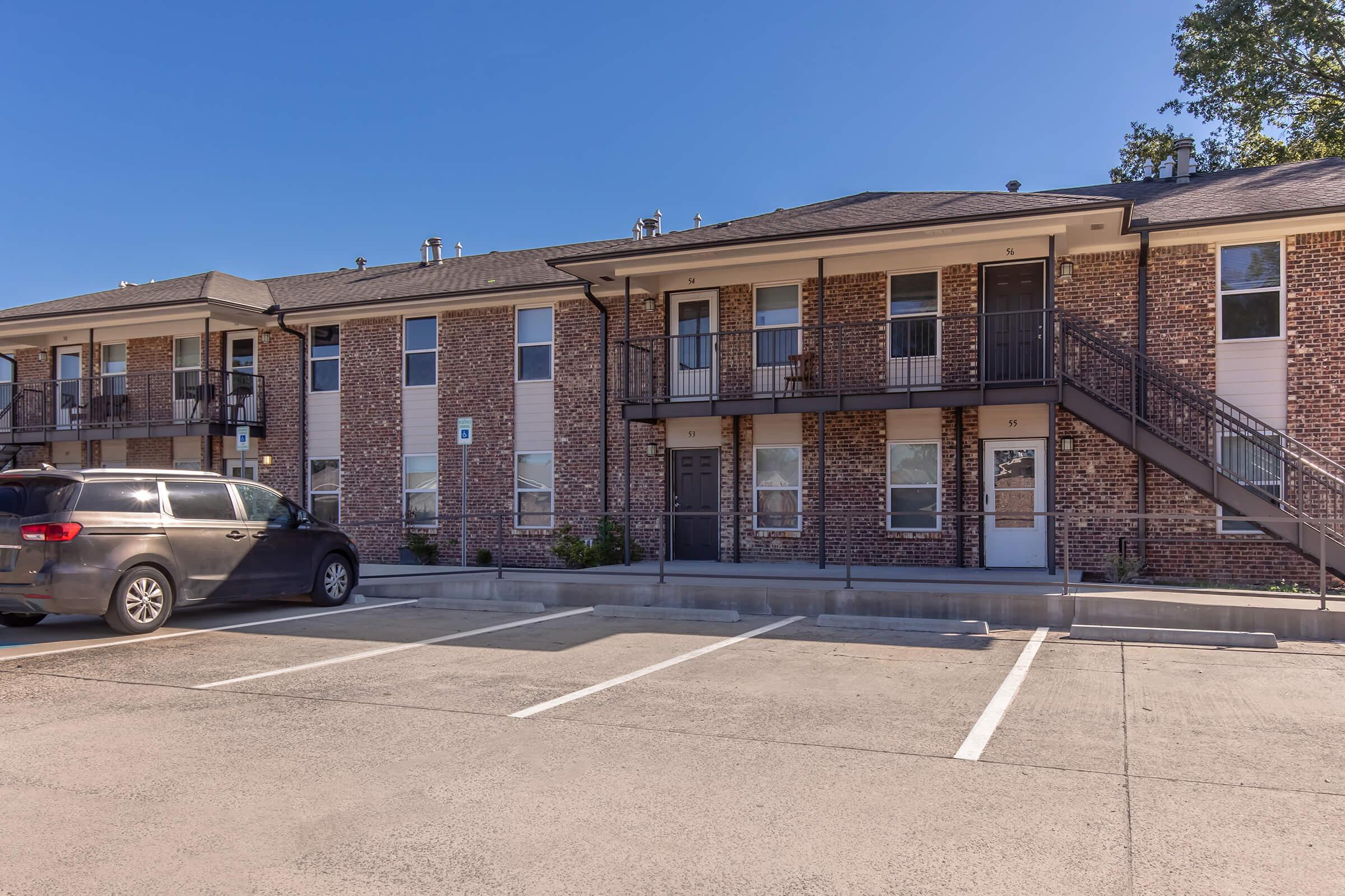 A two-story apartment building made of brick and siding, featuring multiple doors and windows. A parking lot in front has several marked spaces, with a minivan parked nearby. The scene is set under a clear blue sky, showcasing the structure's modern design and well-maintained exterior.