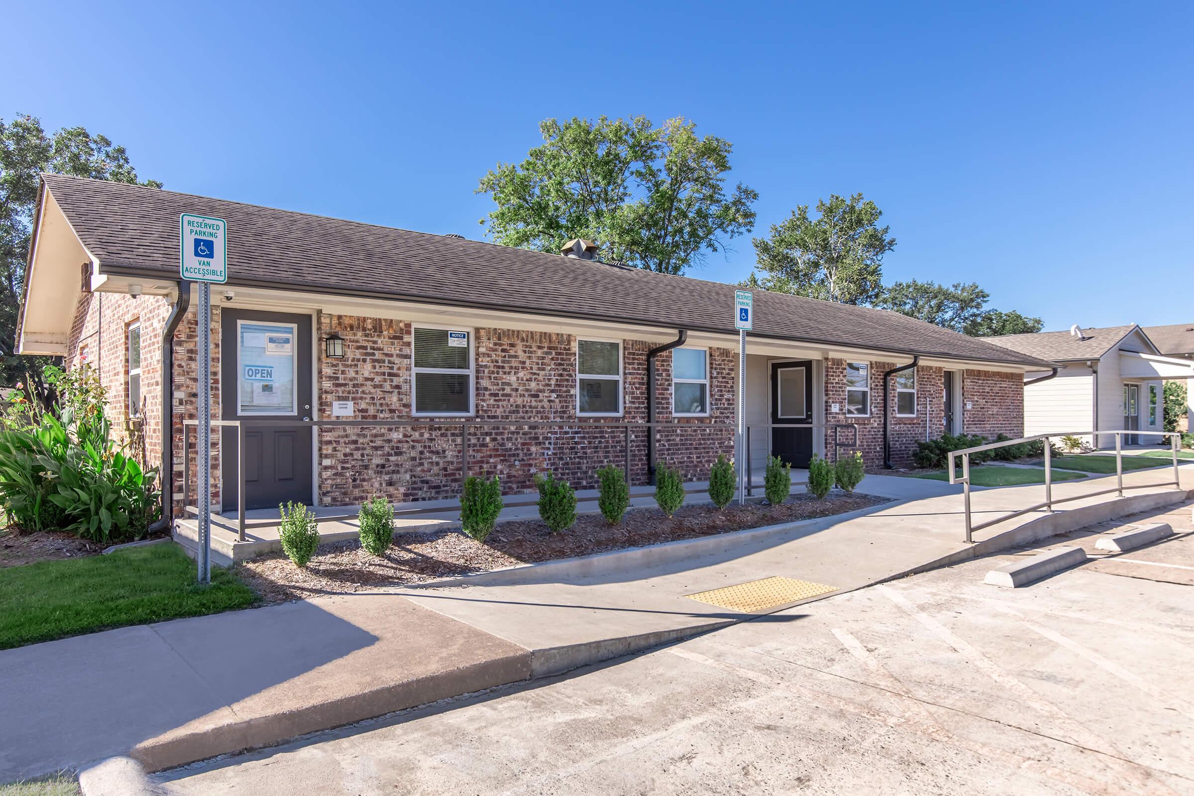 A single-story brick building with a flat roof, featuring a mix of windows and doors. Accessible parking spaces are in front, along with a concrete walkway leading to the entrance. Surrounding landscaping includes small shrubs and trees under clear blue skies.