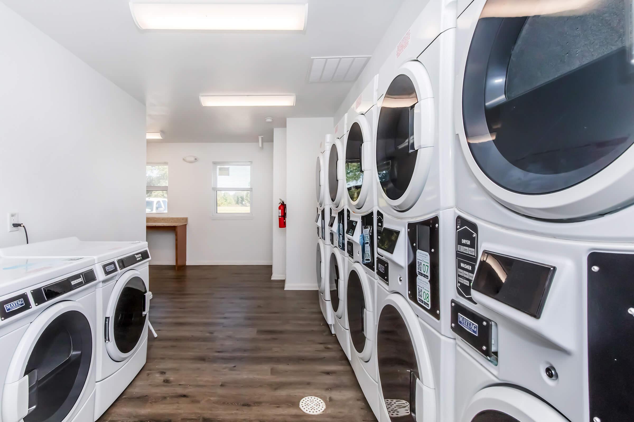 A bright, modern laundry room featuring rows of white washers and dryers with digital displays. The room has a clean, minimalist design with light-colored walls and wood flooring. A large window allows natural light to enter, and a wooden folding table is visible at the far end. A fire extinguisher is mounted on the wall.