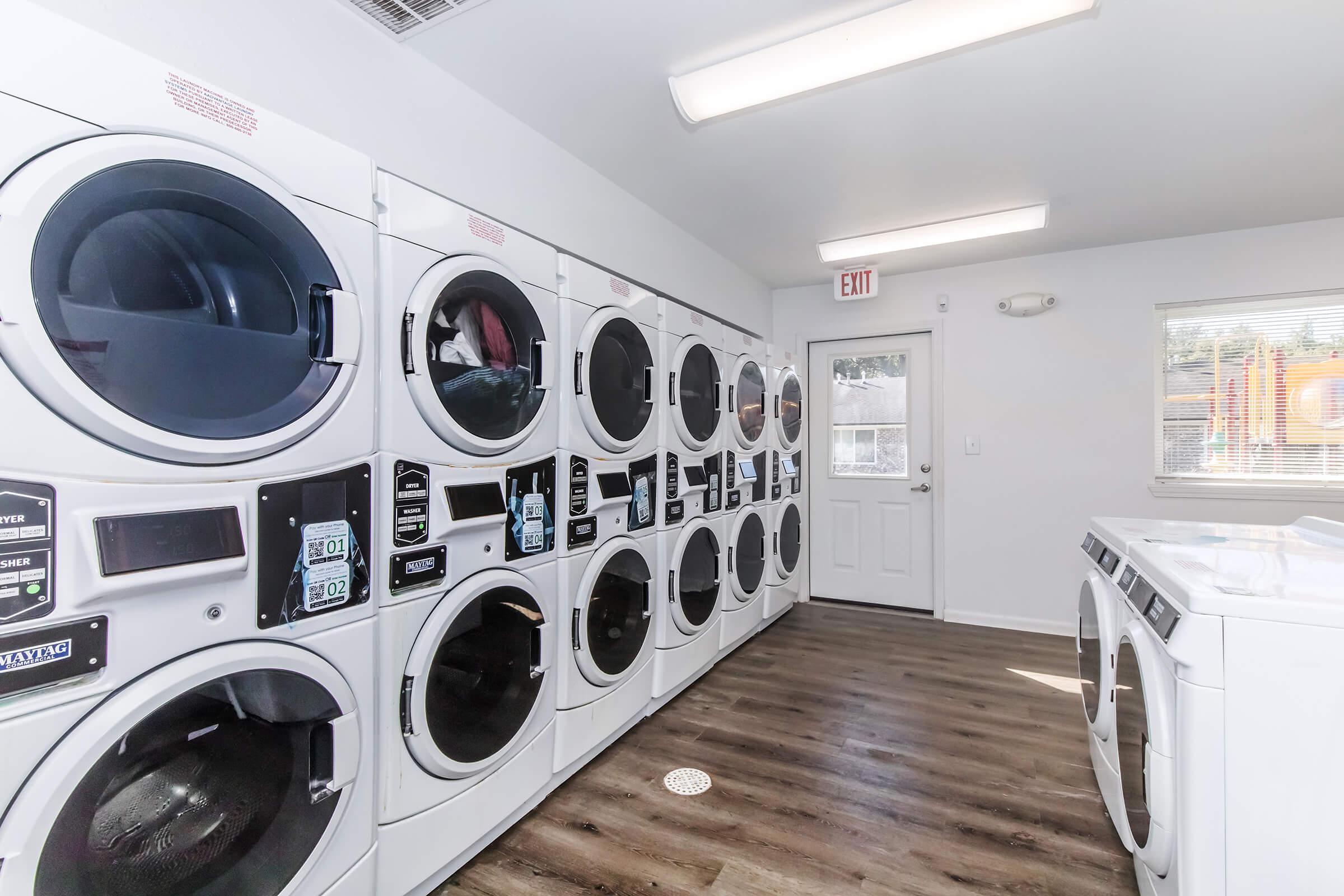 A clean, well-lit laundromat featuring several rows of white washing machines and dryers. The modern appliances have control panels visible, and there's a door leading outside. Bright overhead lights illuminate the space, and a window provides natural light. The flooring is a warm wood finish.