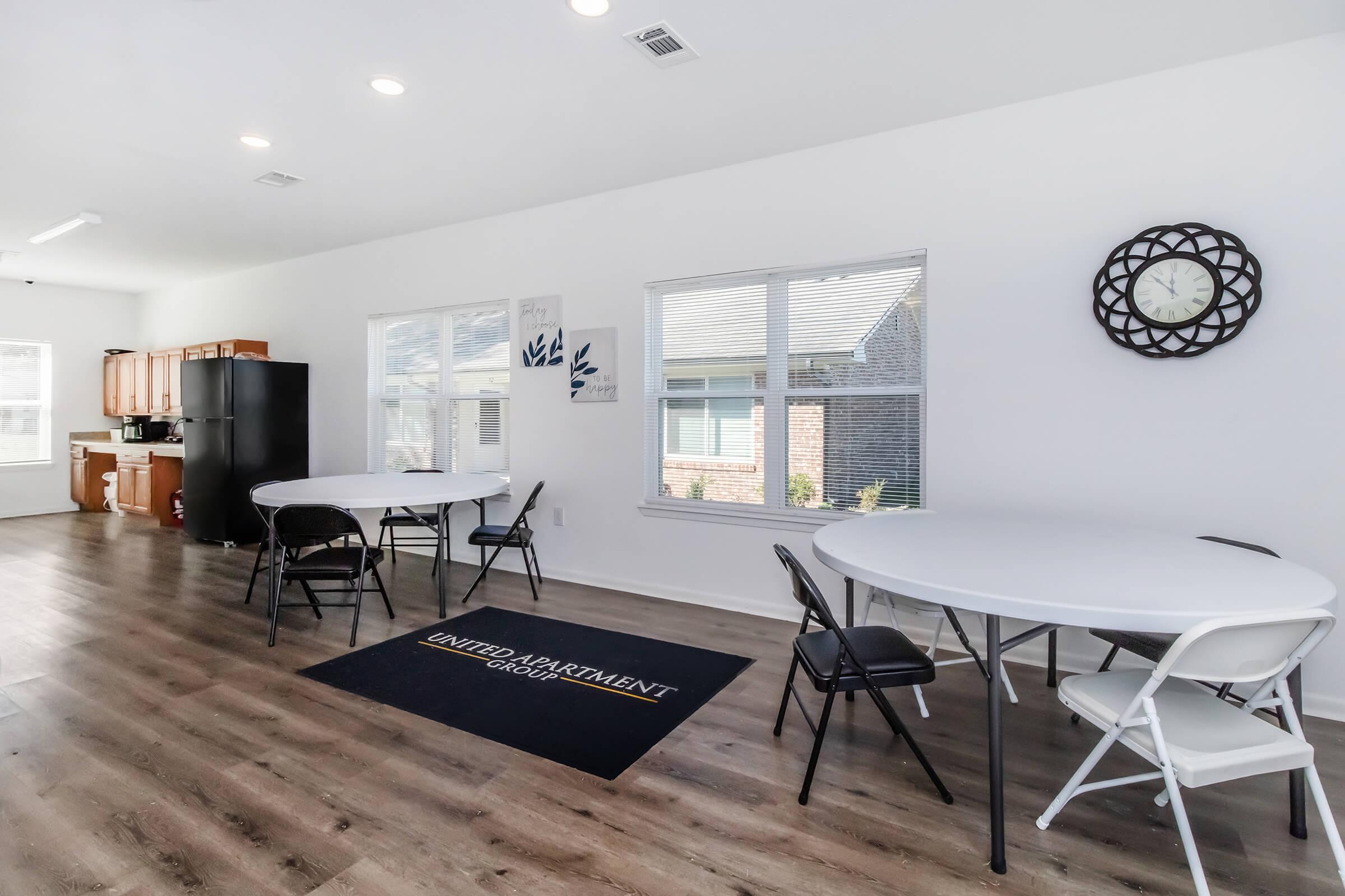 A spacious interior room featuring two round white tables with chairs, a black refrigerator, and large windows allowing natural light. A decorative wall clock is visible, and the floor has a wooden finish. A welcome mat with the text "UNITED APARTMENT" is placed near the entrance.