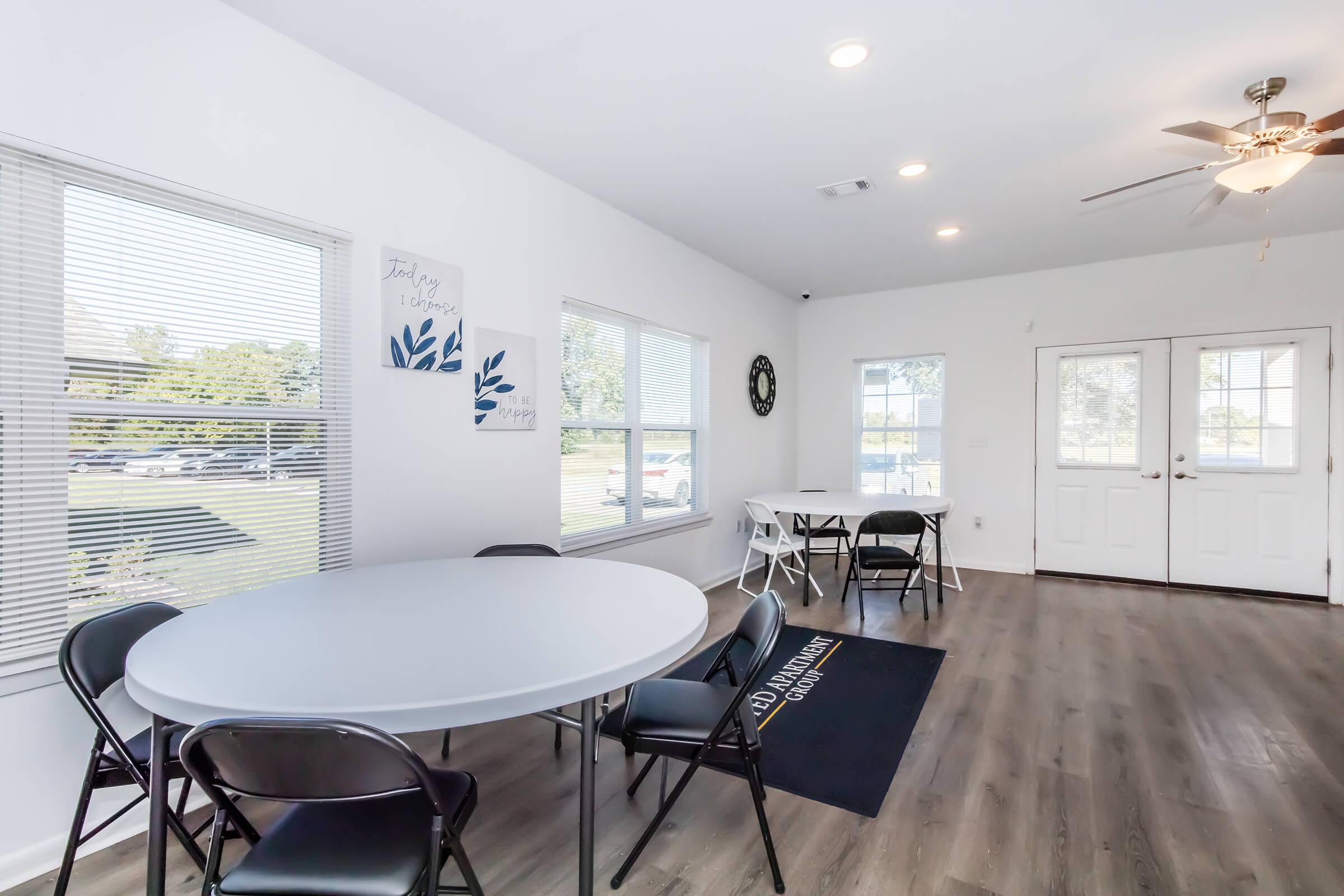 A bright and airy room with large windows, featuring a round table surrounded by black chairs. There are two additional tables, and the space has light-colored walls and flooring. A ceiling fan hangs above, and a decorative wall sign is visible. The entrance is visible with double doors.