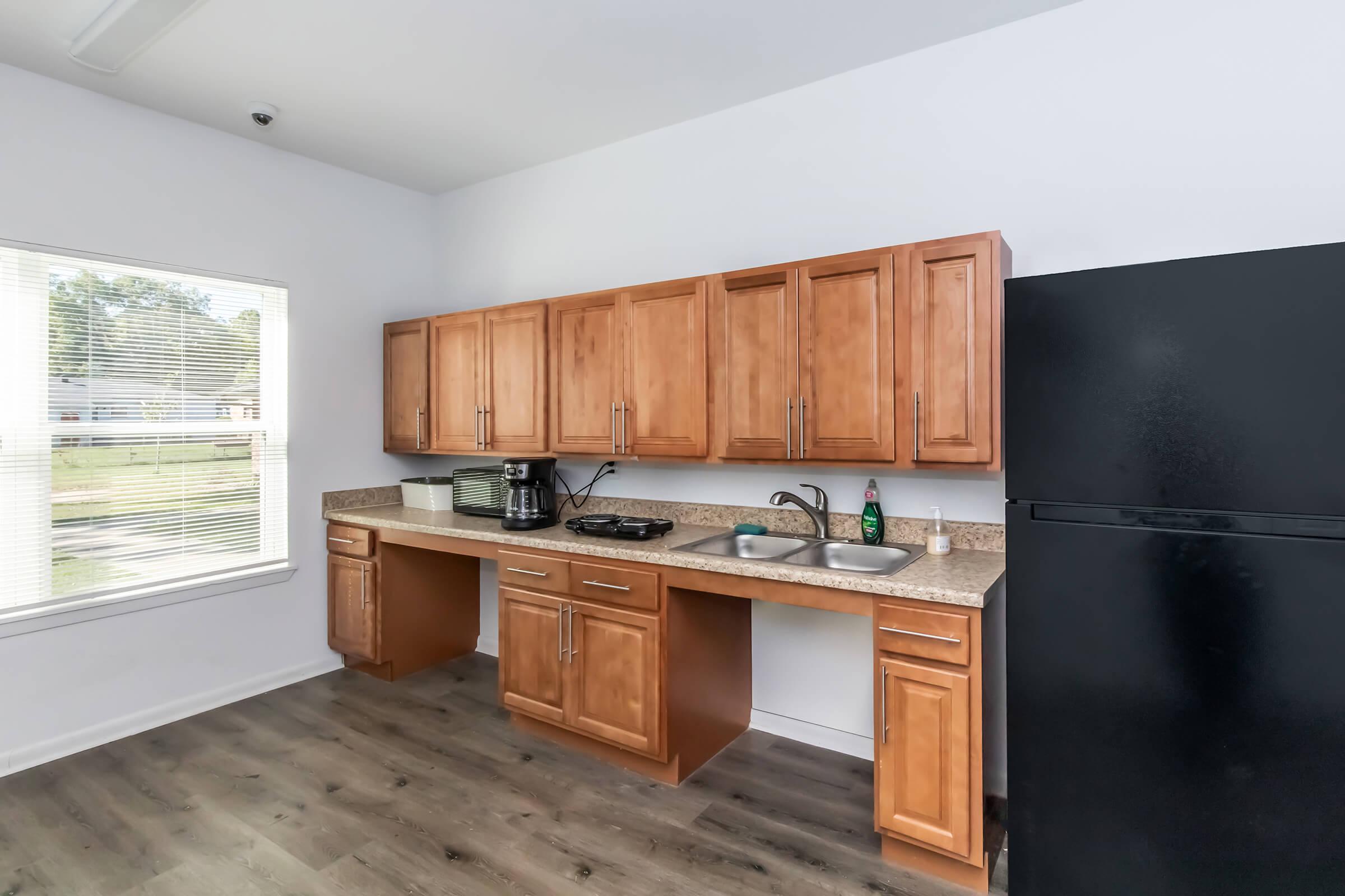 Modern kitchen with wooden cabinetry, a black refrigerator, and a countertop. Features include a sink, stovetop, and a coffee maker. Natural light enters through a window, illuminating the space, which has a light-colored floor.