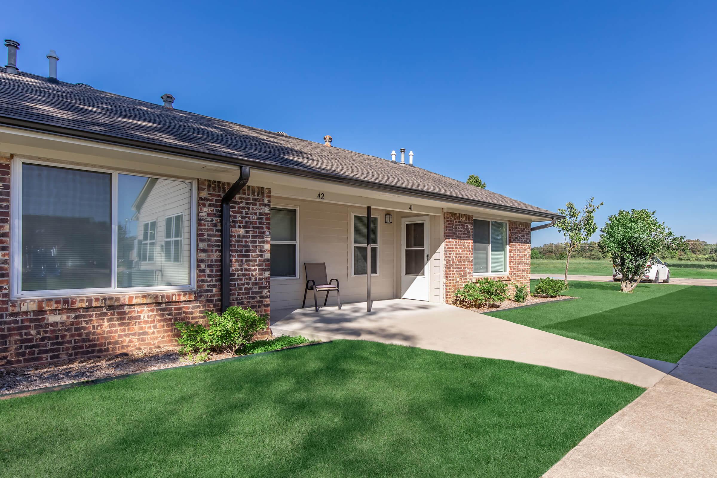 A single-story brick house with a front porch, a chair outside, and well-maintained green lawn. The sky is clear and blue, and there are trees in the background. The driveway is paved, leading to the front entrance.