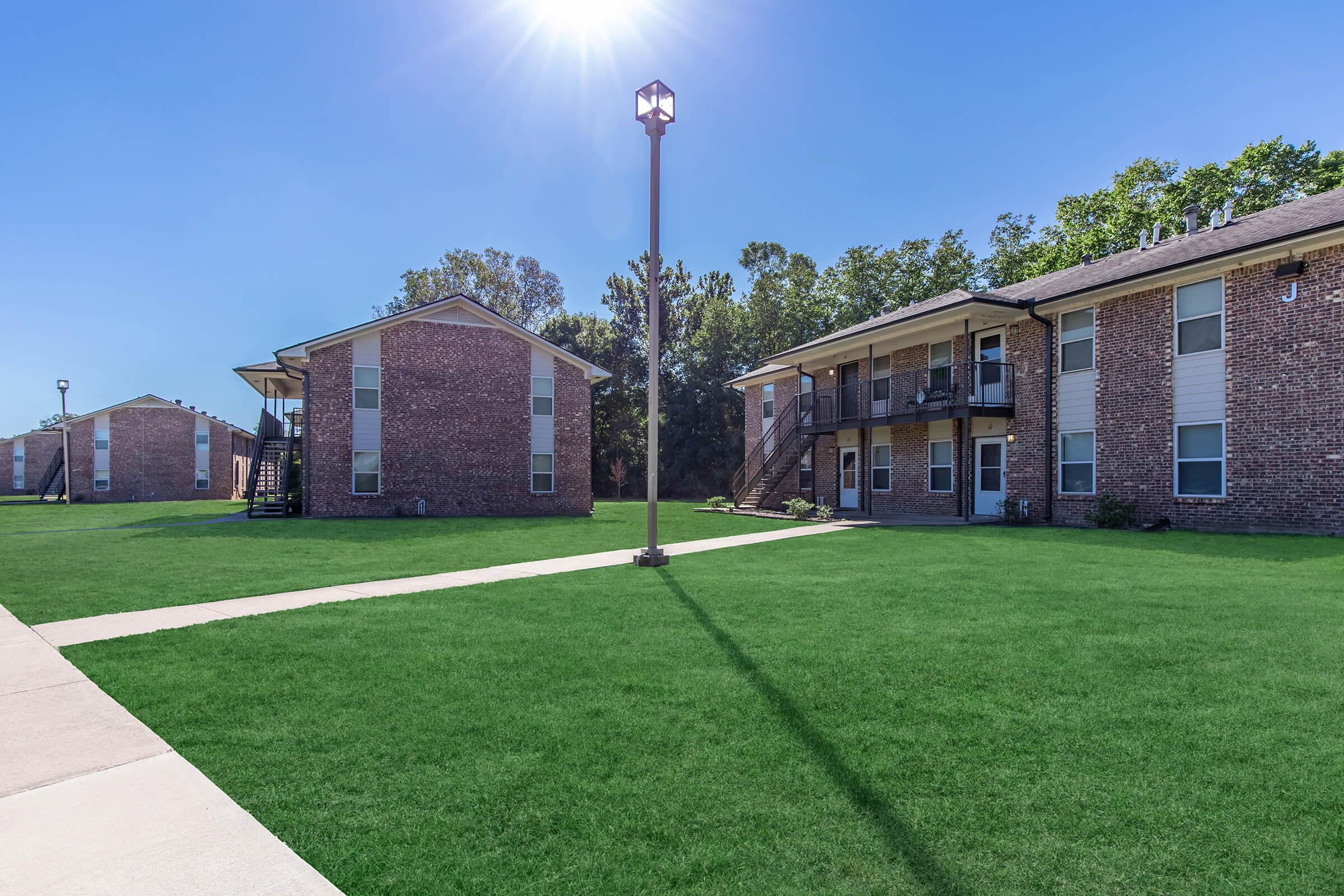 A well-maintained outdoor area featuring two brick apartment buildings arranged around a grassy courtyard. There are walkways leading to the buildings, and a lamp post stands in the center, with a clear blue sky overhead and trees in the background. The setting appears clean and inviting.