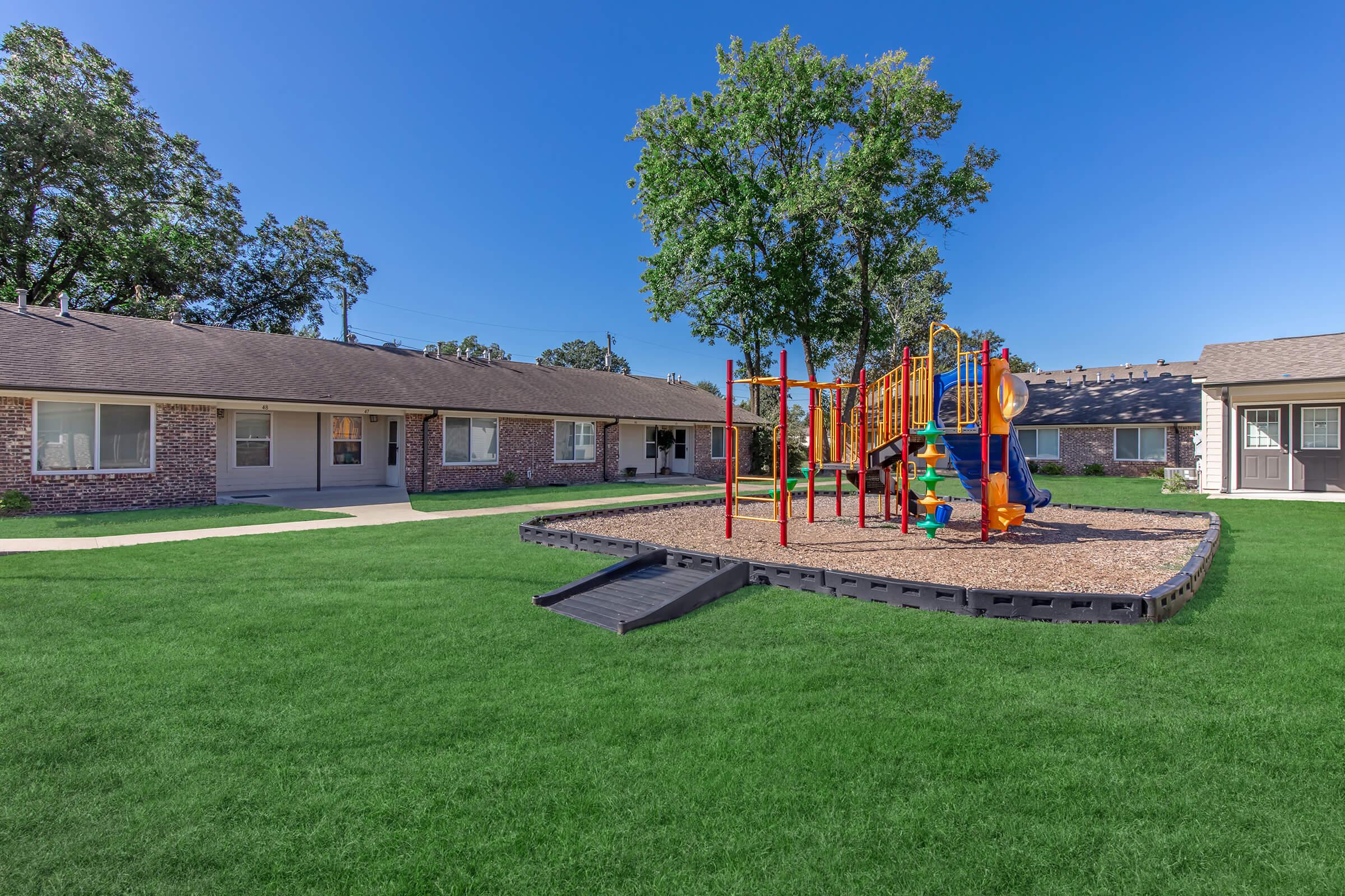A bright, sunny view of a playground with colorful equipment, including slides and climbing structures, situated on a green lawn. In the background, there are two buildings with a brick facade and multiple windows. The sky is clear and blue, enhancing the cheerful atmosphere of the outdoor space.