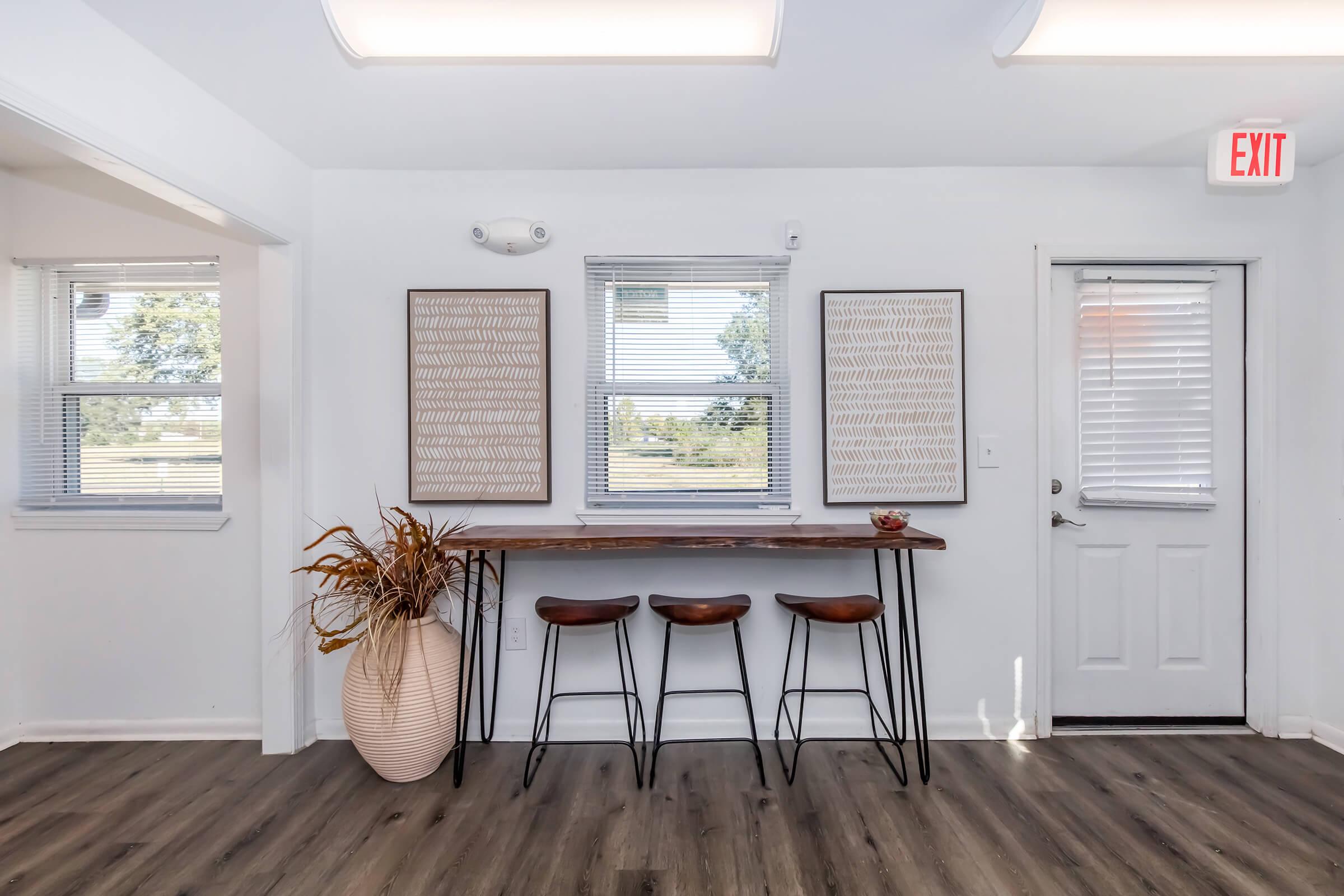 A bright, modern interior featuring a minimalist wooden bar table with three metal stools. On the wall, two decorative framed artworks are displayed. Natural light comes through the windows, and a potted plant sits on the table. A door with a safety exit sign is visible on the right.