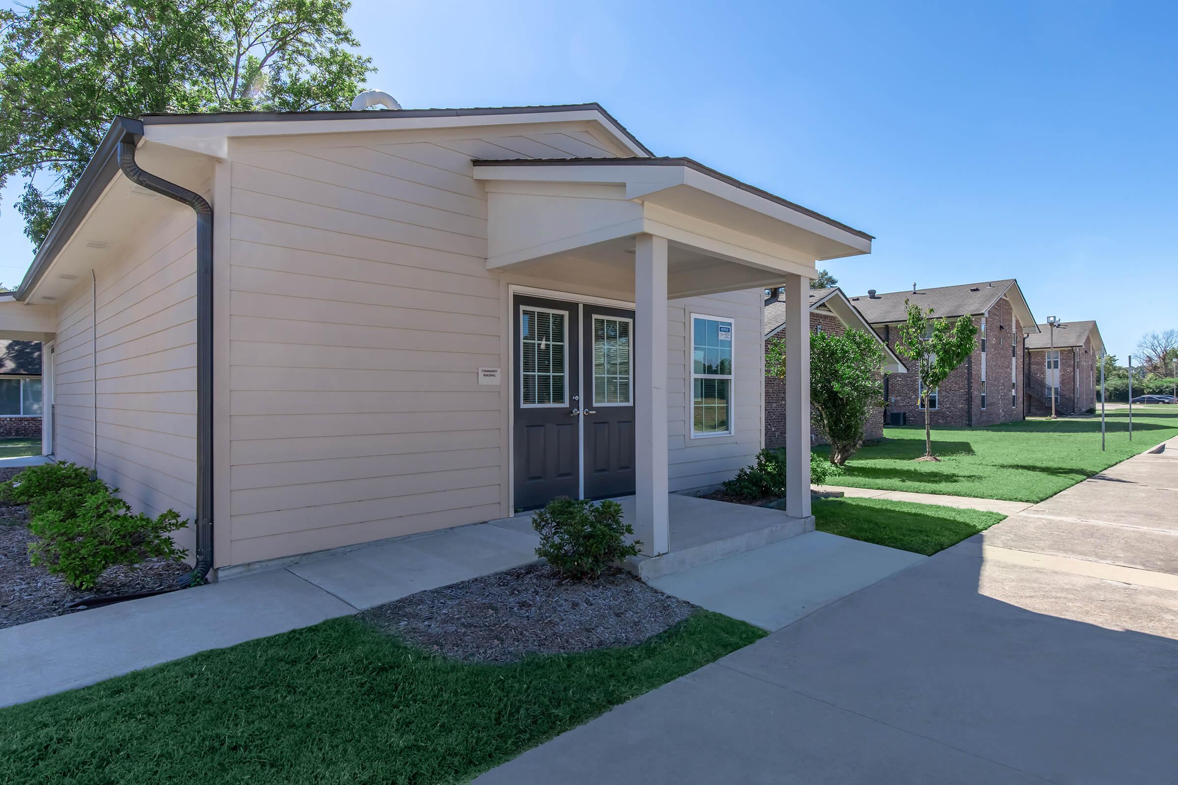 A light-colored residential building with a covered porch, two front doors, and large windows. The landscape features well-manicured green lawns and trees. In the background, additional brick buildings are visible under a clear blue sky. The pathway is made of concrete.