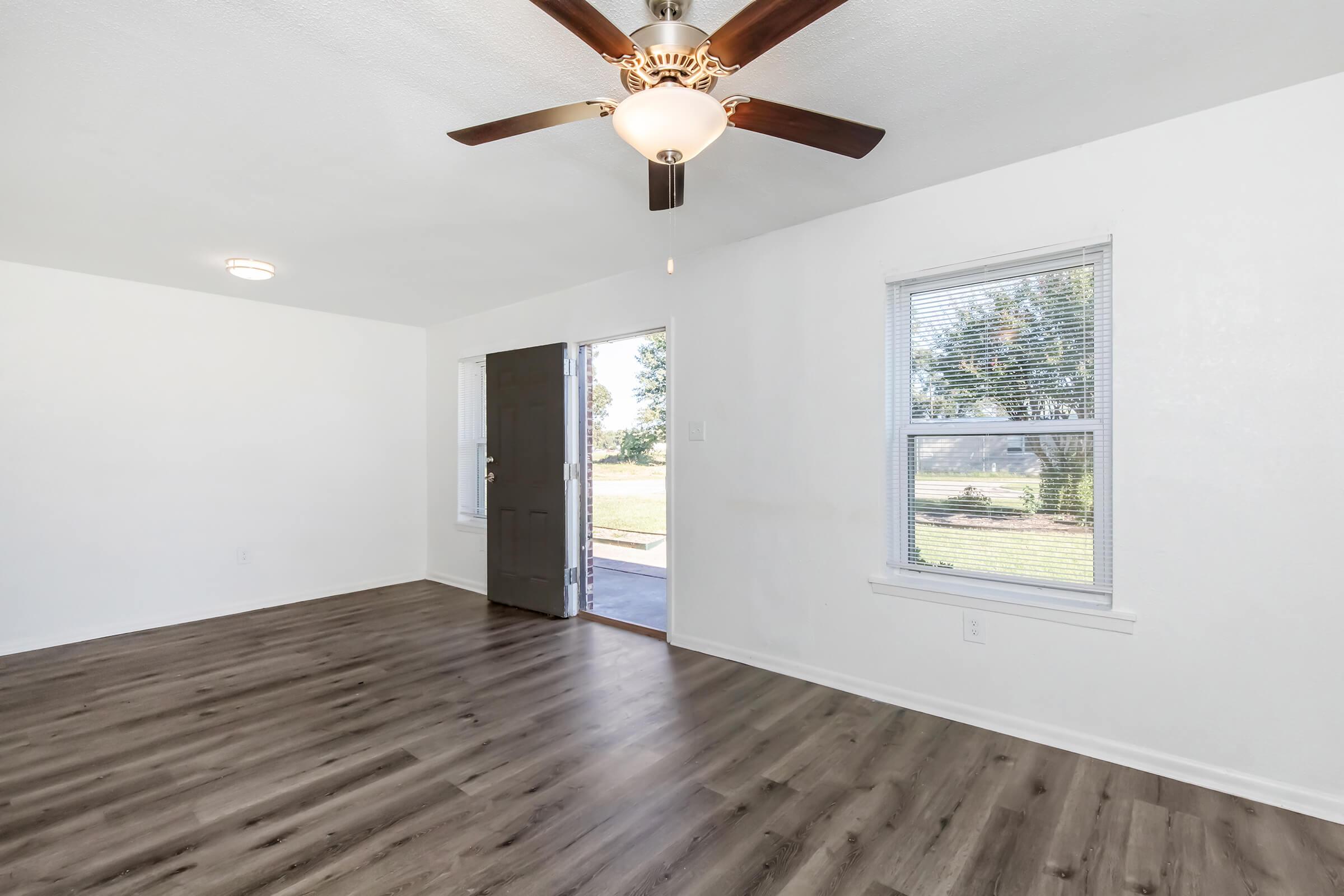 Interior view of a bright, empty room featuring a light-colored wall, a ceiling fan with light fixture, and a front door leading outside. Two windows provide natural light, and the floor is covered with wood-like laminate. The space is ready for furniture and decoration.