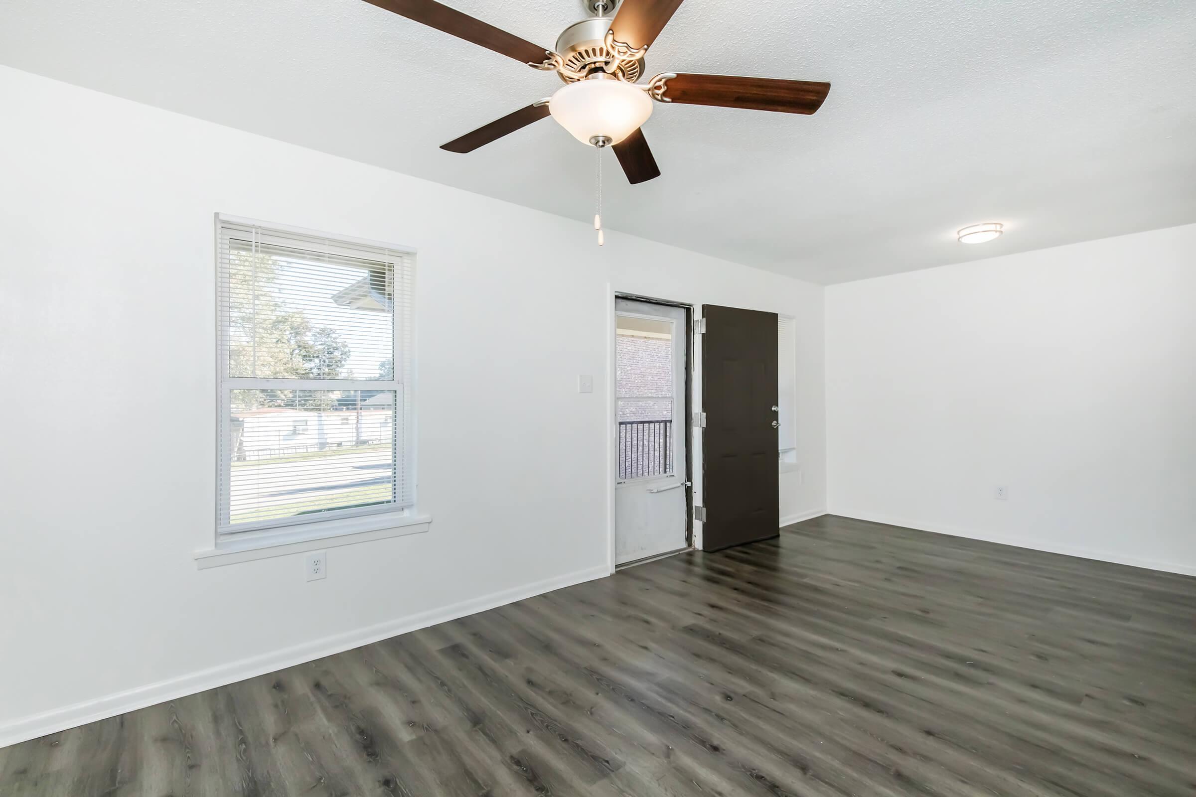 A spacious, well-lit room with light-colored walls and a ceiling fan. There is a window with blinds allowing natural light, and a door leading outside. The floor is covered with modern, dark laminate wood flooring.