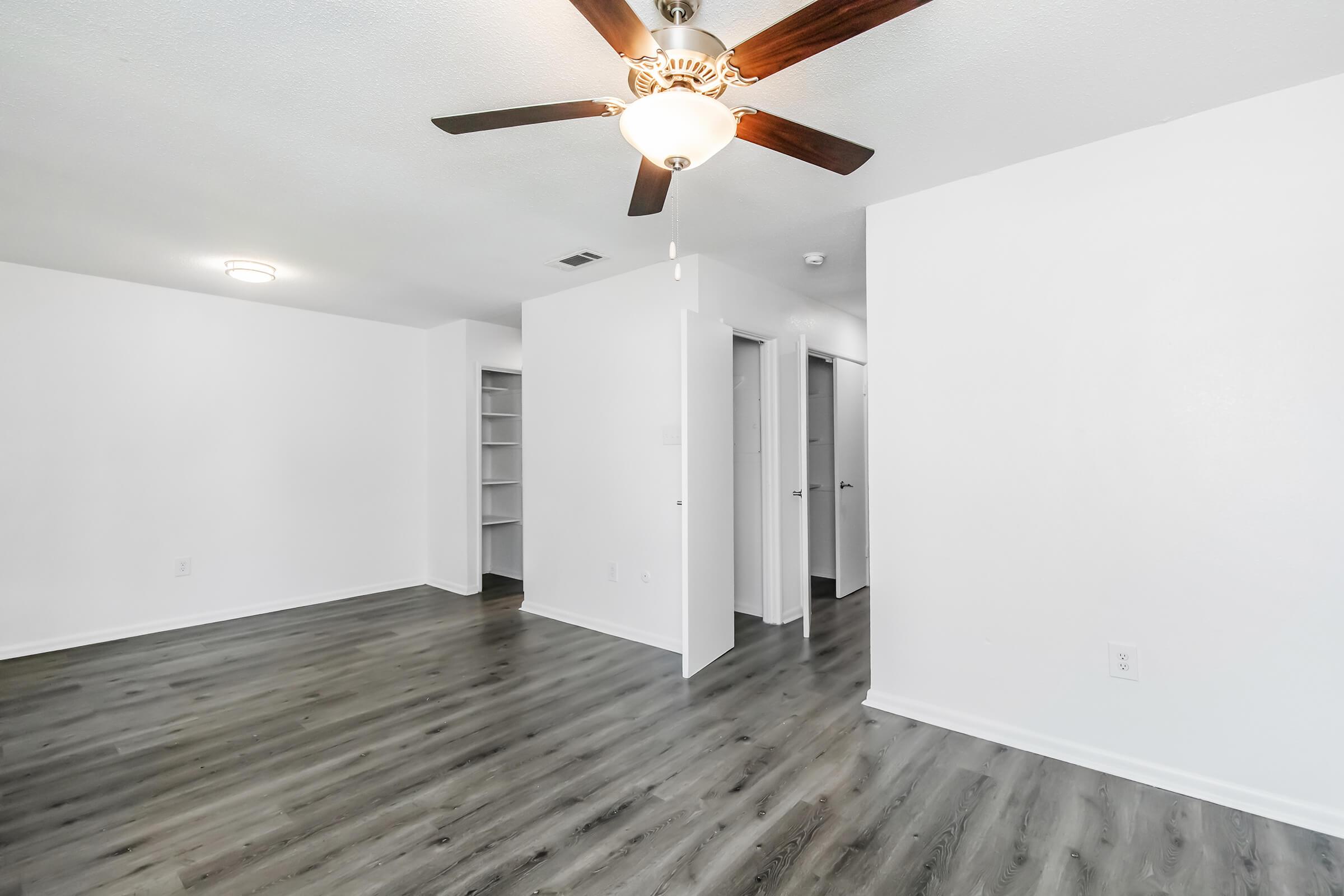 A spacious, well-lit room featuring a ceiling fan, light-colored walls, and gray laminate flooring. There is a closet on one side and two doorways leading to adjacent rooms, creating an open and inviting layout.
