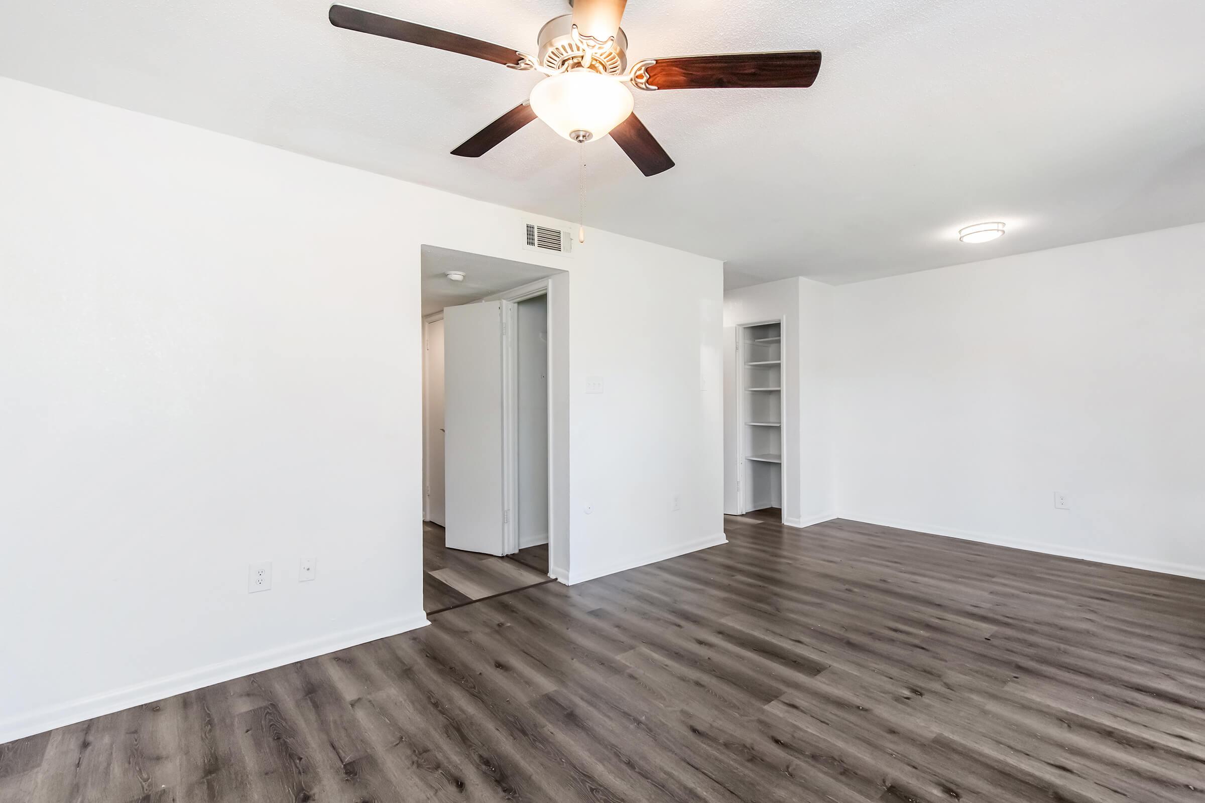 A spacious room with light-colored walls and a ceiling fan, featuring laminate flooring. To the left, there is an open doorway leading to a hallway. On the right side, a built-in shelf can be seen against the wall, creating a minimalist and bright atmosphere.