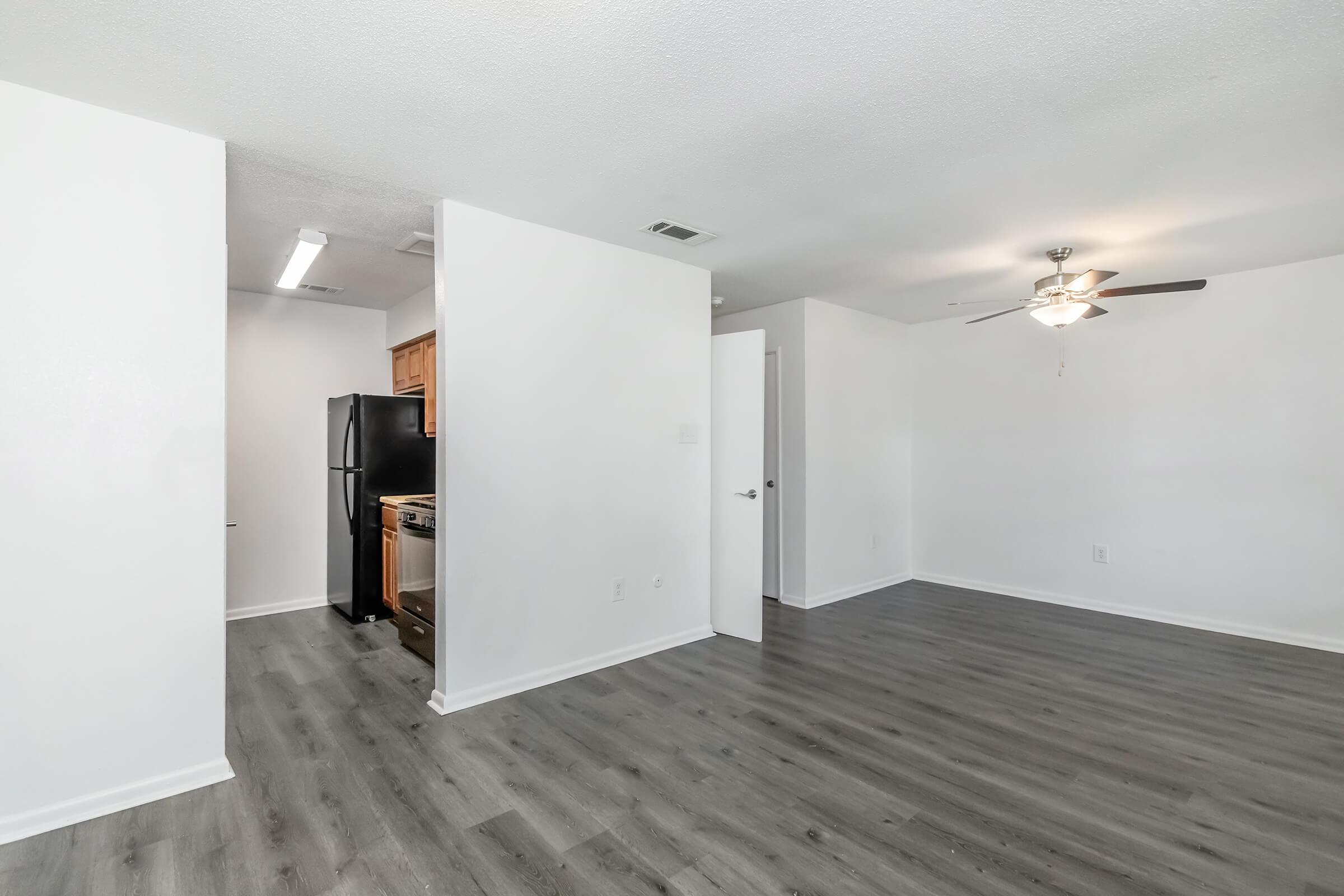 A modern, open-concept living space featuring light-colored walls, laminate flooring, and a ceiling fan. To the left, there is a kitchen area with dark appliances, including a refrigerator and stove, visible through an archway. The room has ample natural light and space for furniture arrangements.