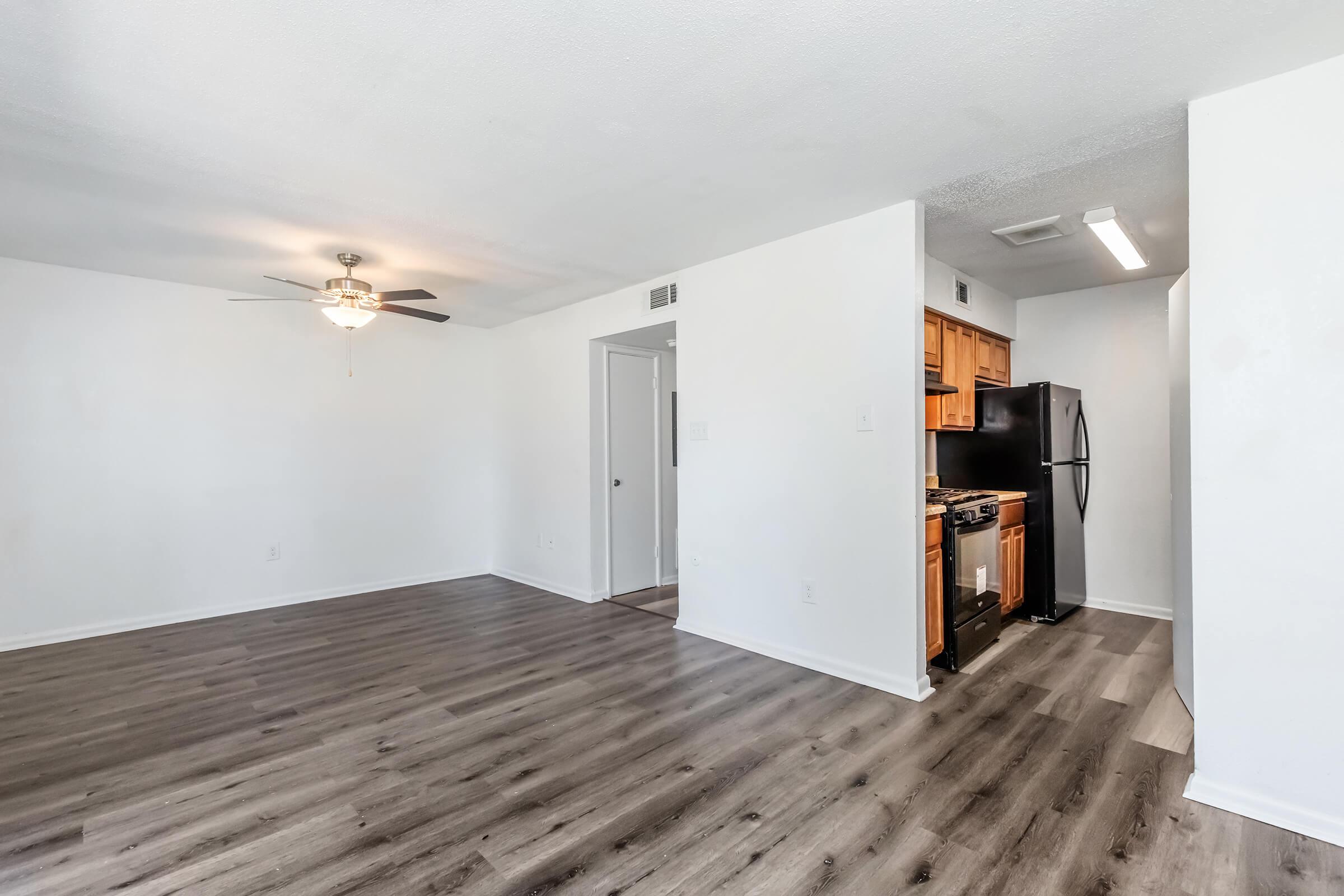 Spacious open living area featuring a ceiling fan, modern laminate flooring, and a kitchen area with wooden cabinets and stainless steel appliances. The walls are painted white, creating a bright and inviting atmosphere. A doorway leads to a bedroom or bathroom off to one side.