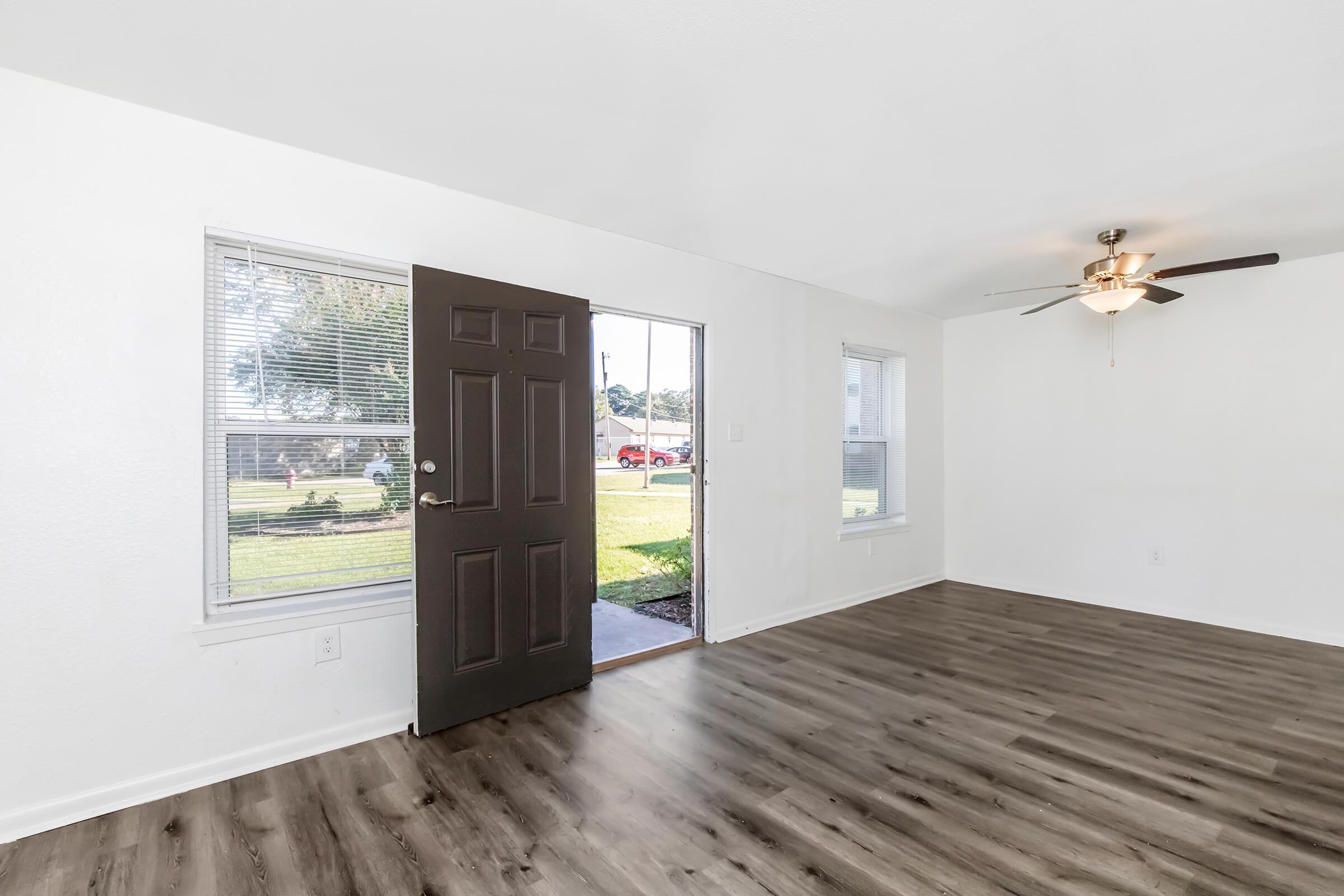 A bright, empty room with a dark front door and two windows allowing natural light to enter. The flooring is a light-colored laminate, and a ceiling fan is visible above. Outside the window, there's a view of green grass and a sidewalk. The walls are painted white, creating a fresh, modern look.