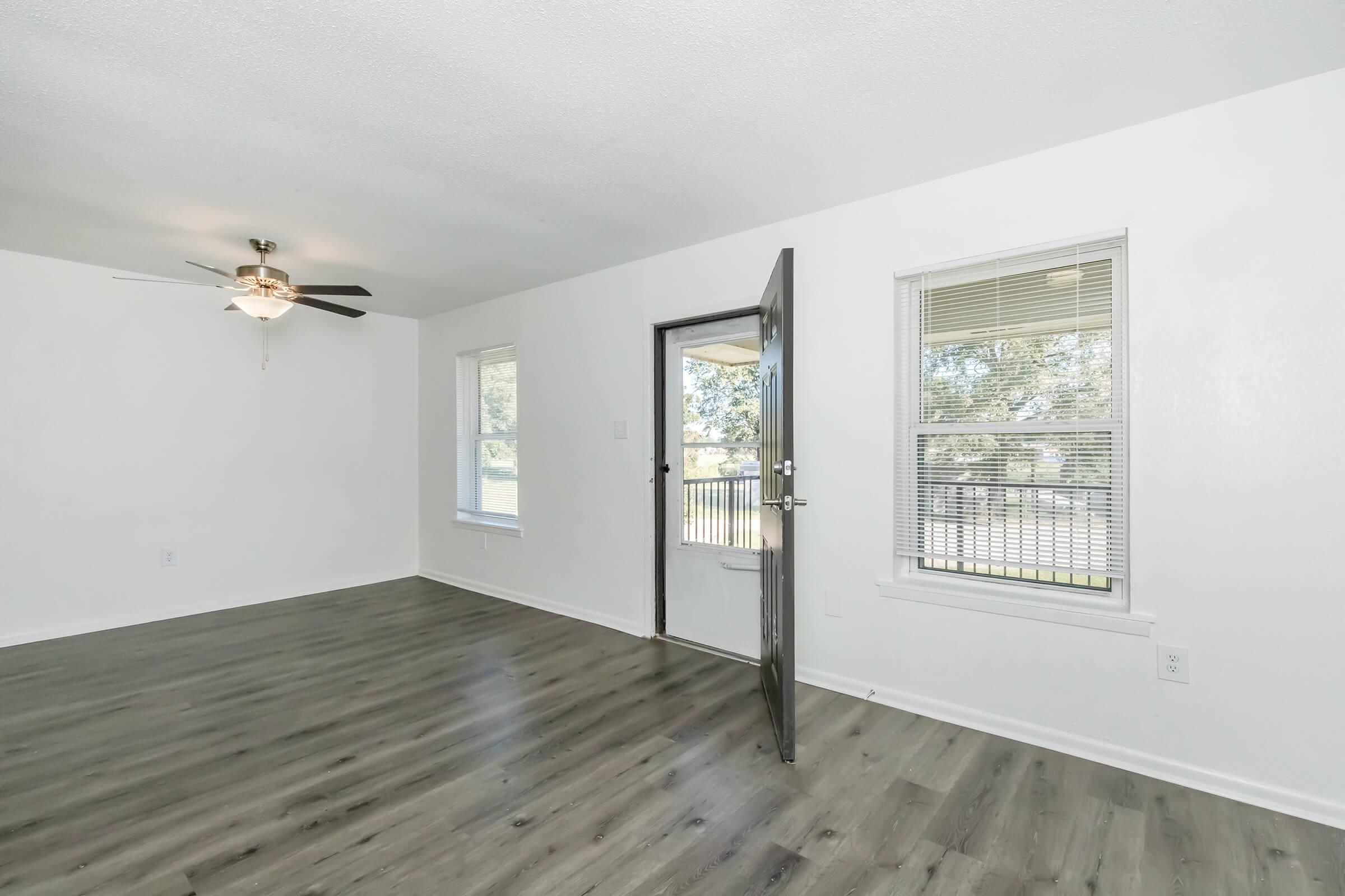 Spacious living room with light-colored walls and wood-look flooring. A ceiling fan hangs from the ceiling, and a door is open to a patio. There are two windows allowing natural light to enter the space, enhancing the bright and airy atmosphere.