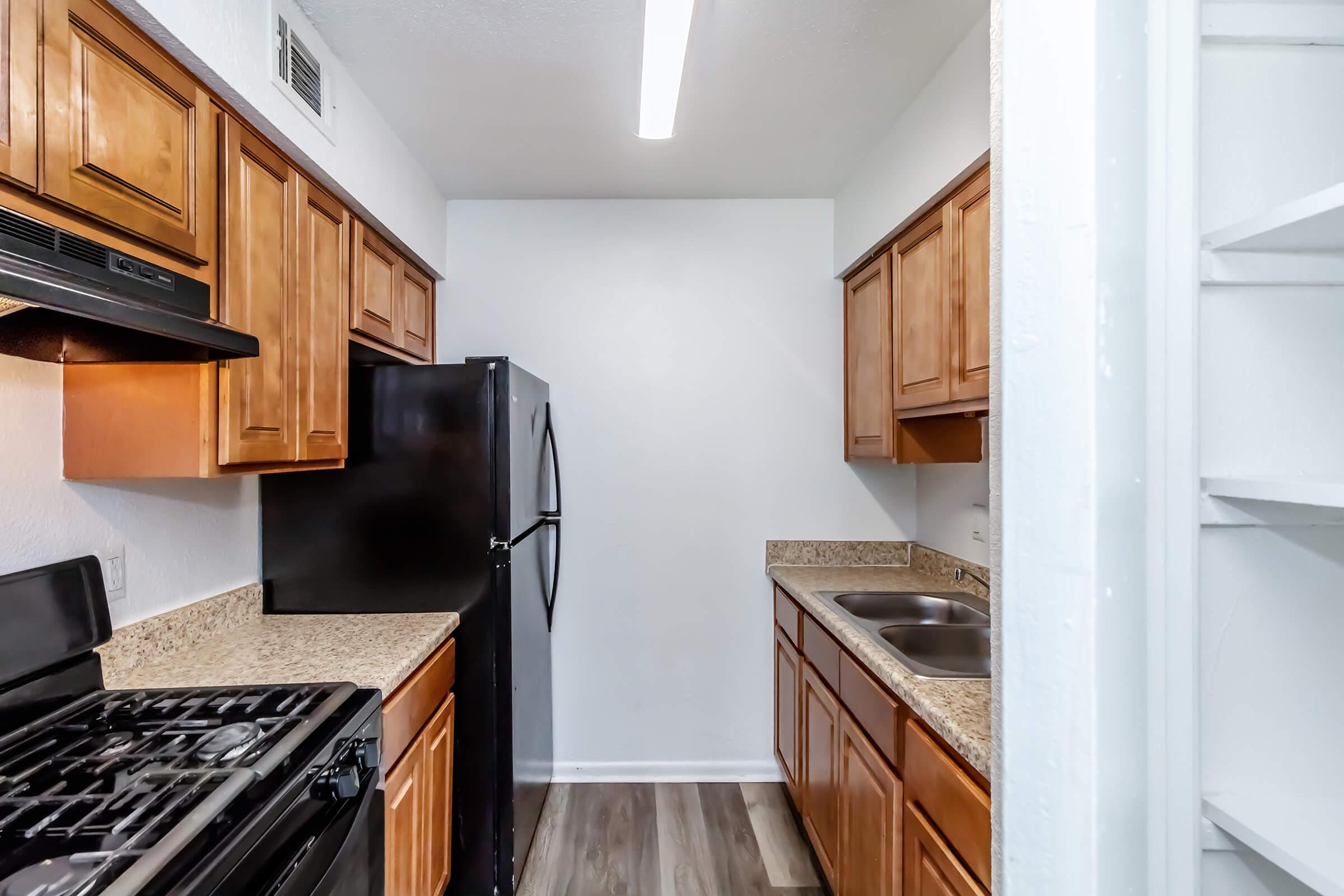 Modern kitchen featuring wooden cabinets, a black refrigerator, a gas stove, and dual sinks. The countertops are a light granite, and the flooring is a wood-style laminate. Bright lighting from a ceiling fixture illuminates the space, which has white walls and a simple layout.