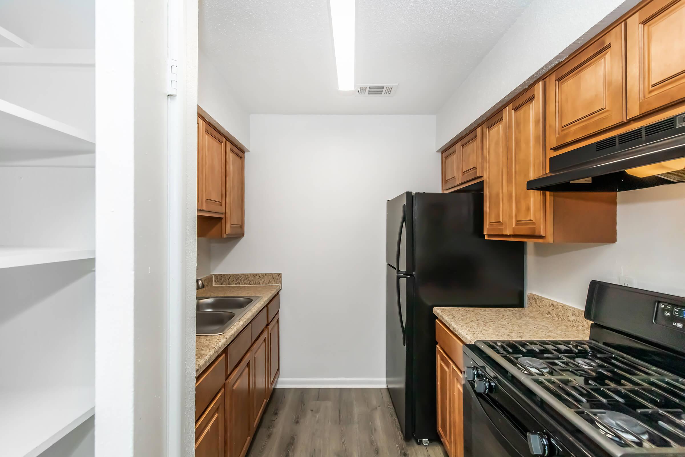 Modern kitchen featuring wooden cabinets, black appliances, and granite countertops. Includes a double sink, a refrigerator, and an oven with a gas stove. A pantry area is visible on the left, and the flooring is a light-colored wood laminate.