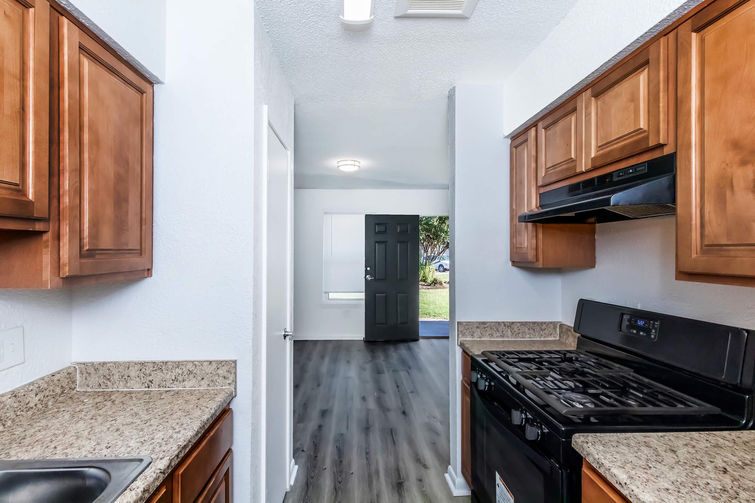 View from the kitchen showcasing wooden cabinets, a granite countertop, and a black stove. The hallway leads to a door at the end, opening to a well-lit living area with a large window and modern flooring.