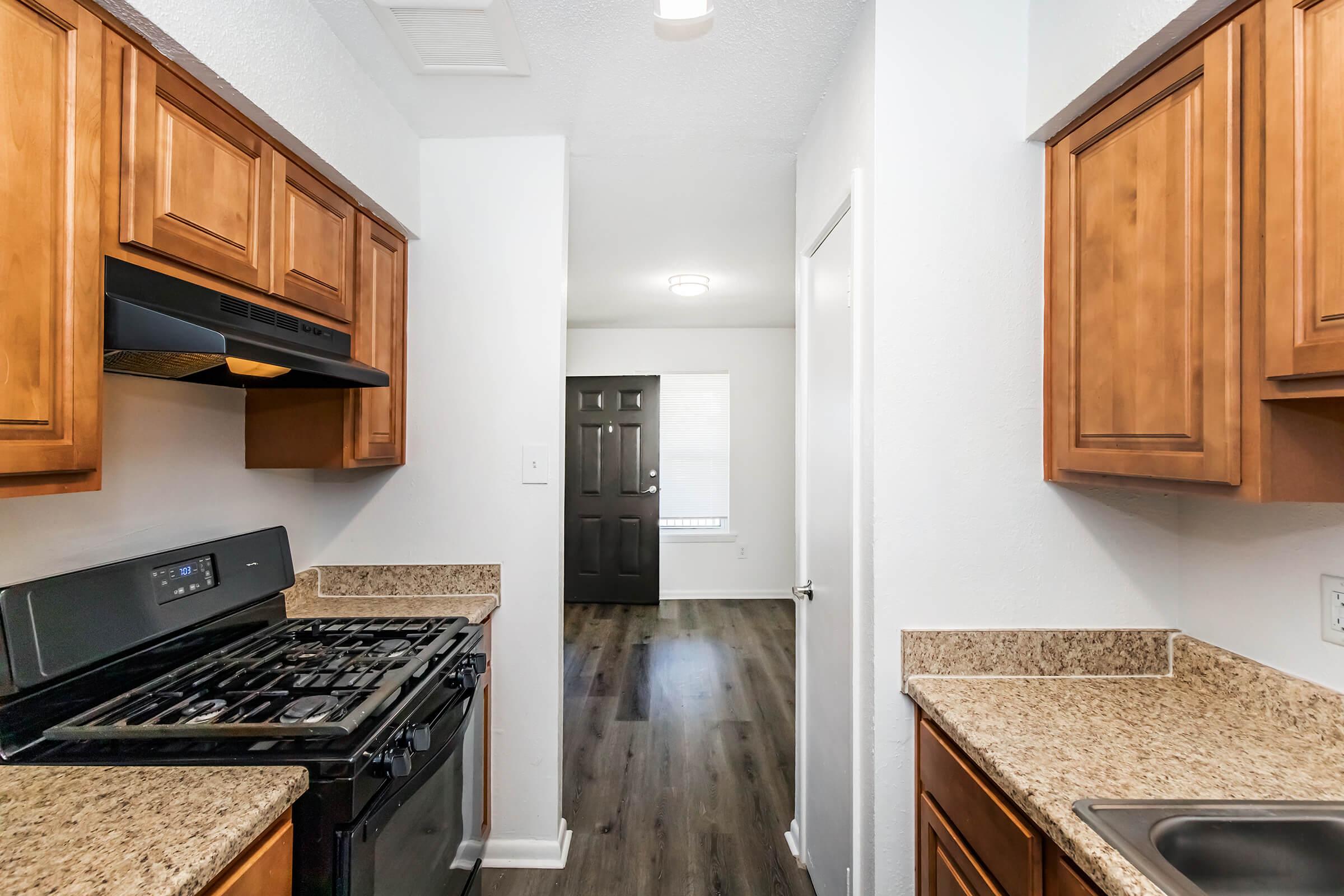 A modern kitchen featuring wooden cabinetry, granite countertops, a black gas stove, and a sink. The view extends into a well-lit living area with a dark-colored door and a window, showcasing a clean and inviting home environment.