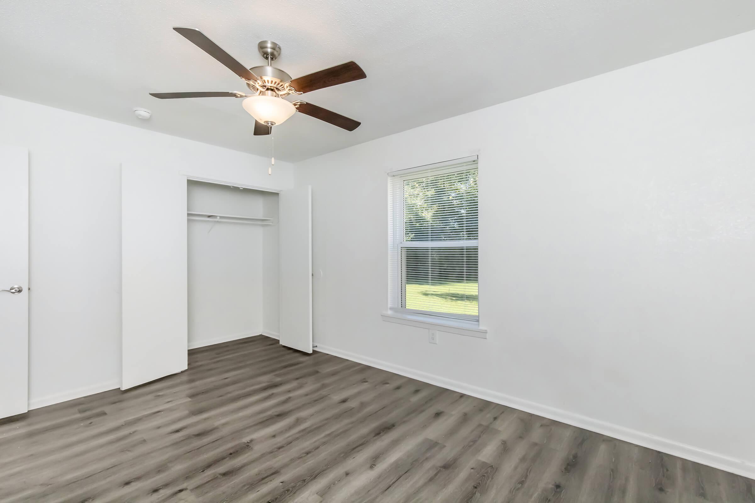 A well-lit room featuring a ceiling fan, light wood-look flooring, and white walls. There are two closet doors on one side and a window with blinds providing natural light and a view of greenery outside.
