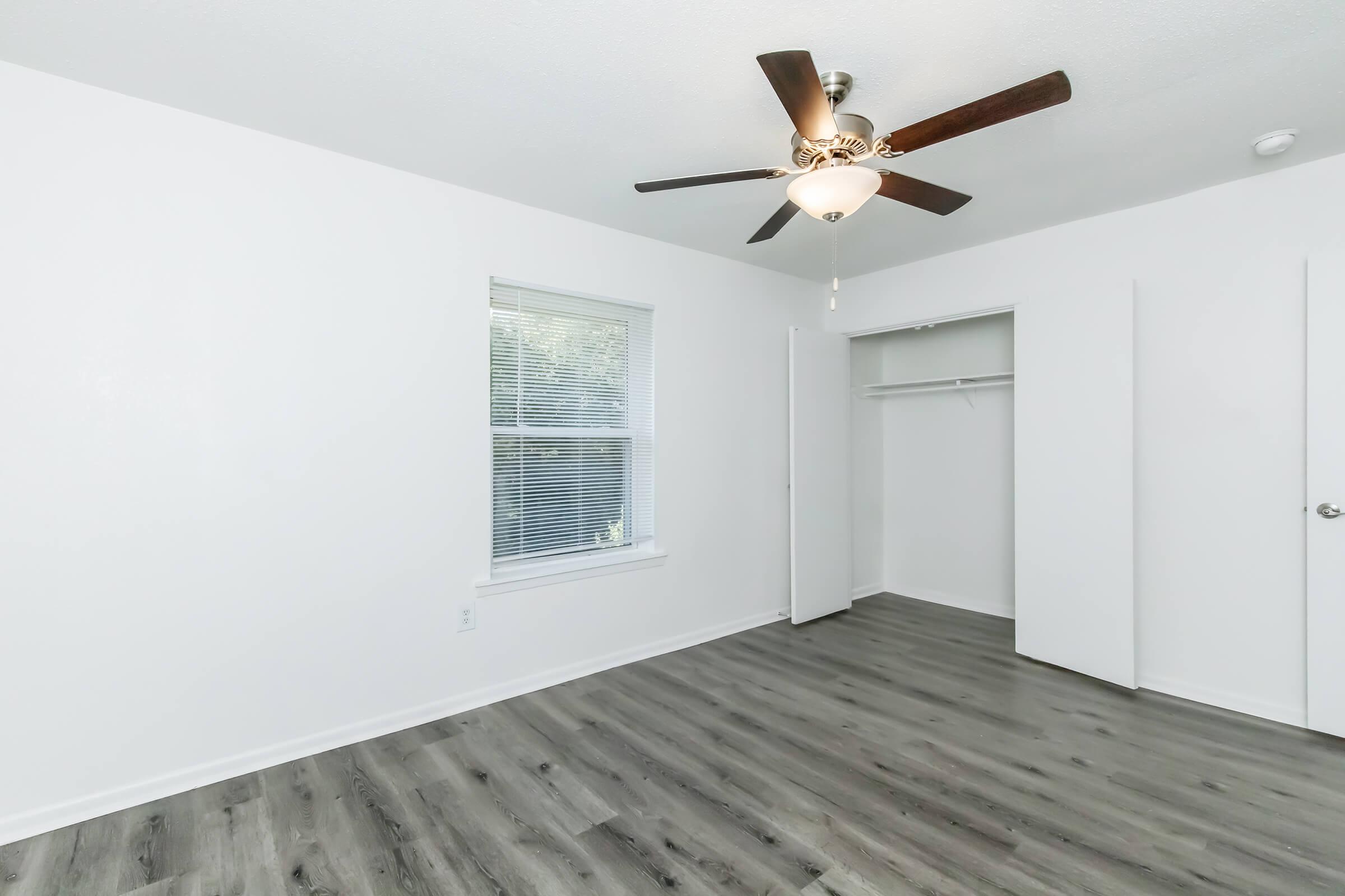 A bright, unfurnished bedroom featuring light-colored walls, a ceiling fan with a light fixture, and a window allowing natural light. The room has dark wood-like flooring and a closet with sliding doors, providing an open and spacious feel.