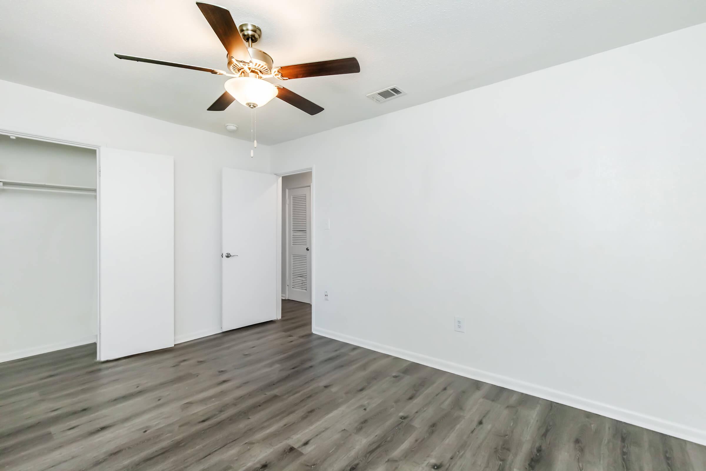 Empty room featuring light-colored walls and a ceiling fan with wooden blades. The floor is a dark wood laminate. There are two closed doors: one leading to a closet and the other likely to a hallway or additional space. The room is well-lit with natural light.