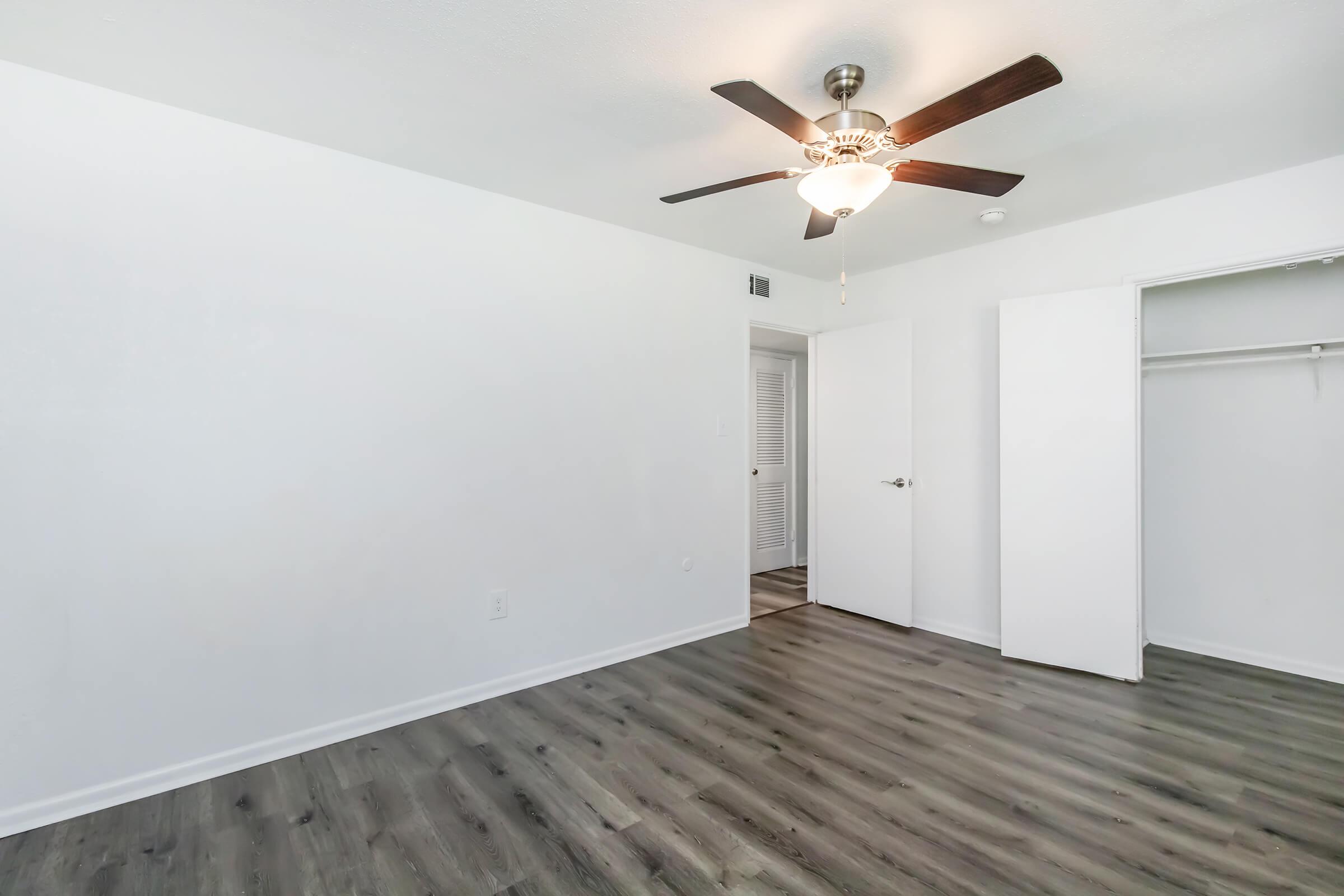 A clean, empty room with light-colored walls, a ceiling fan, and a small closet with sliding doors. The floor is a dark wood laminate, providing a modern look. Bright natural light illuminates the space, giving it an airy feel.