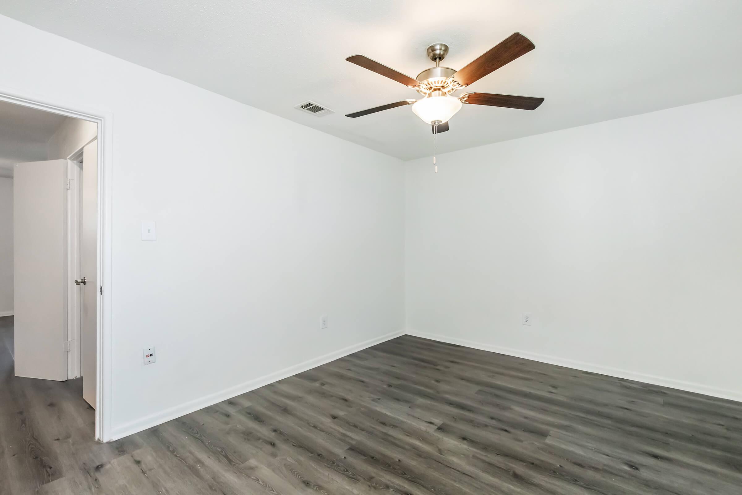 A clean, empty room featuring light gray walls and a ceiling fan with wooden blades. The floor is covered in laminate wood, and there are doors leading to other areas. The space is well-lit and simple, ideal for various uses or decorating.