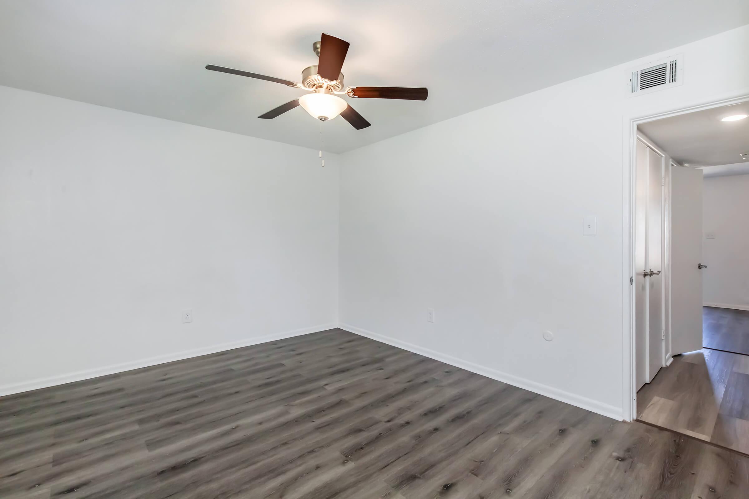 An empty room with light gray walls and a ceiling fan. The floor features laminate wood, and there are doors leading to other areas. The lighting is bright, enhancing the spacious feel of the room.