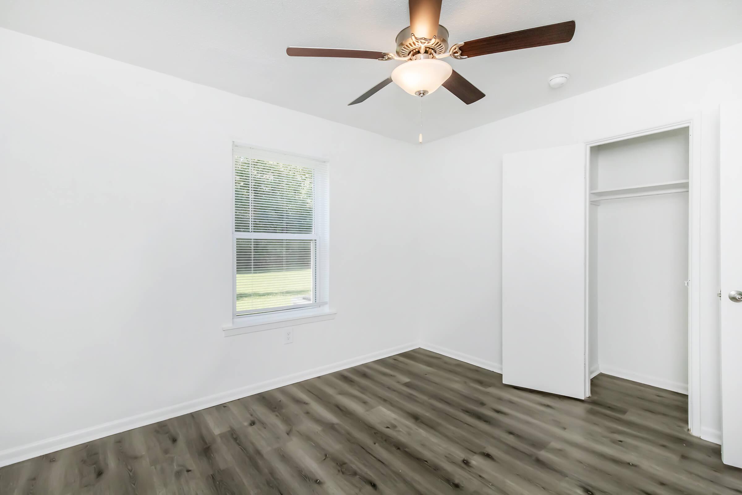 A bright, empty room featuring a ceiling fan with a light fixture, a window allowing natural light, and a closet with sliding doors. The walls are painted white, and the floor has a modern wood-like laminate finish.