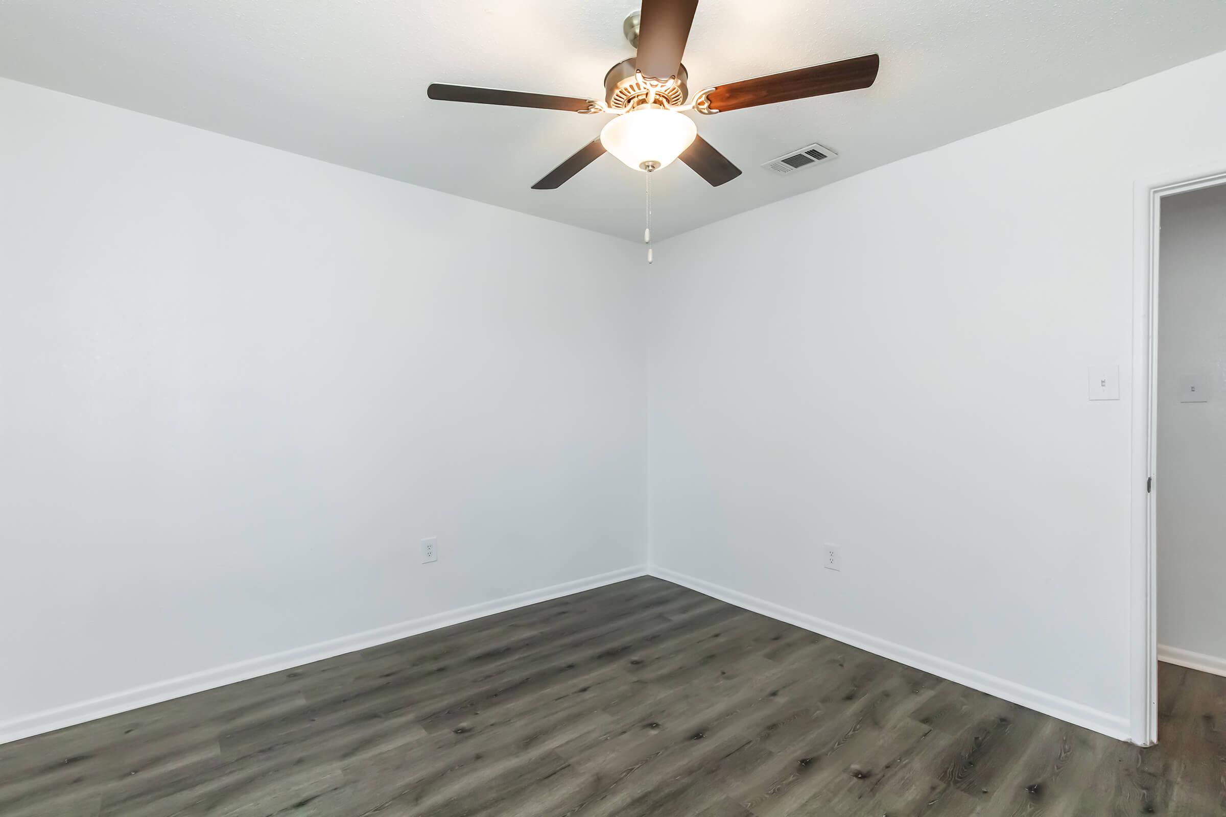 A bright, empty room featuring a ceiling fan with light fixture. The walls are painted white, and the floor is a modern, dark wood laminate. A door on the right leads to another space, and the overall atmosphere is clean and airy, suitable for various uses.